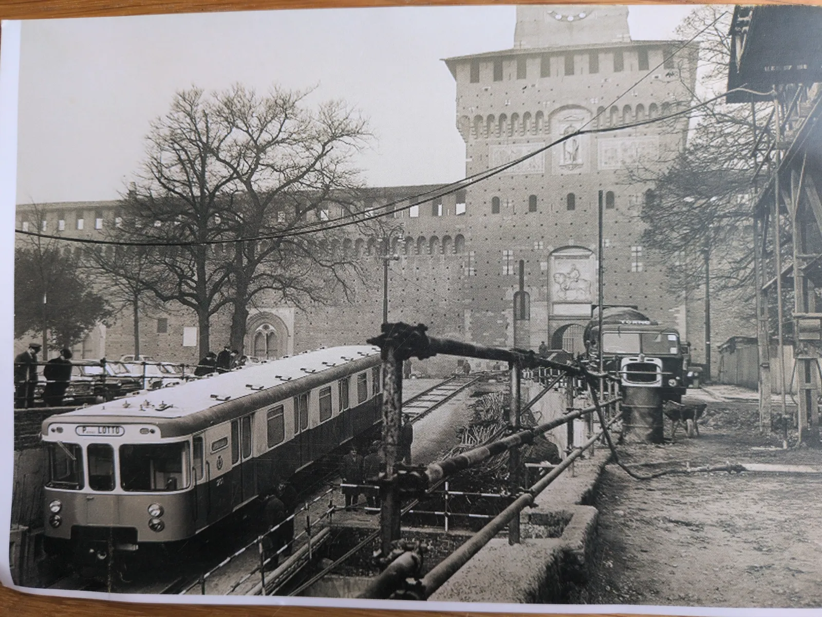 Ein Schwarz-Weiß-Foto zeigt einen modernen Zug auf Gleisen im Vordergrund, vor der Kulisse einer imposanten historischen Burg, während rechts eine Baustelle mit einem Lastwagen zu sehen ist.