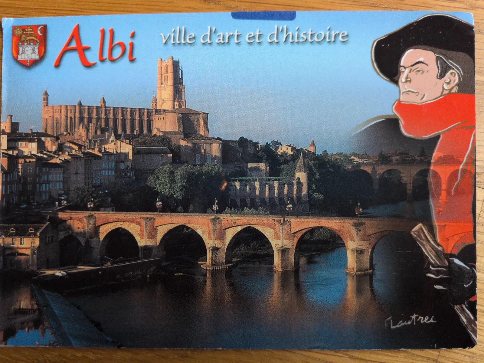 Eine Postkarte zeigt die Stadt Albi mit ihrer großen Kathedrale und einer alten Steinbrücke über einen Fluss, überlagert von einem stilisierten Porträt von Toulouse-Lautrec.