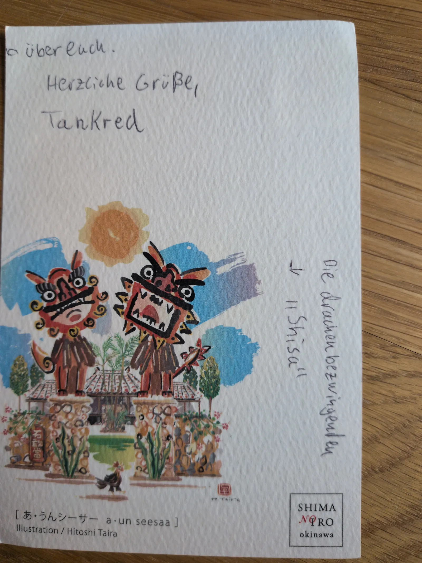 Eine Postkarte zeigt eine bunte Illustration von zwei Shisa-Wächterfiguren aus Okinawa und handschriftliche Grüße.