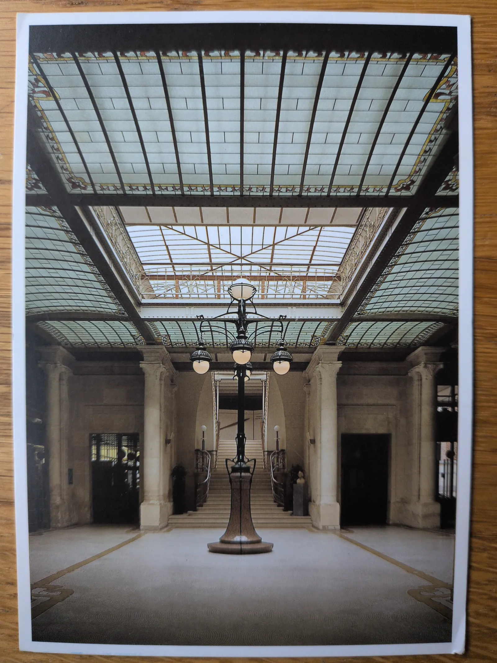 Eine symmetrische Aufnahme einer prunkvollen Halle mit einer großen Standleuchte in der Mitte, einer breiten Treppe im Hintergrund und einem kunstvollen Glasdach.