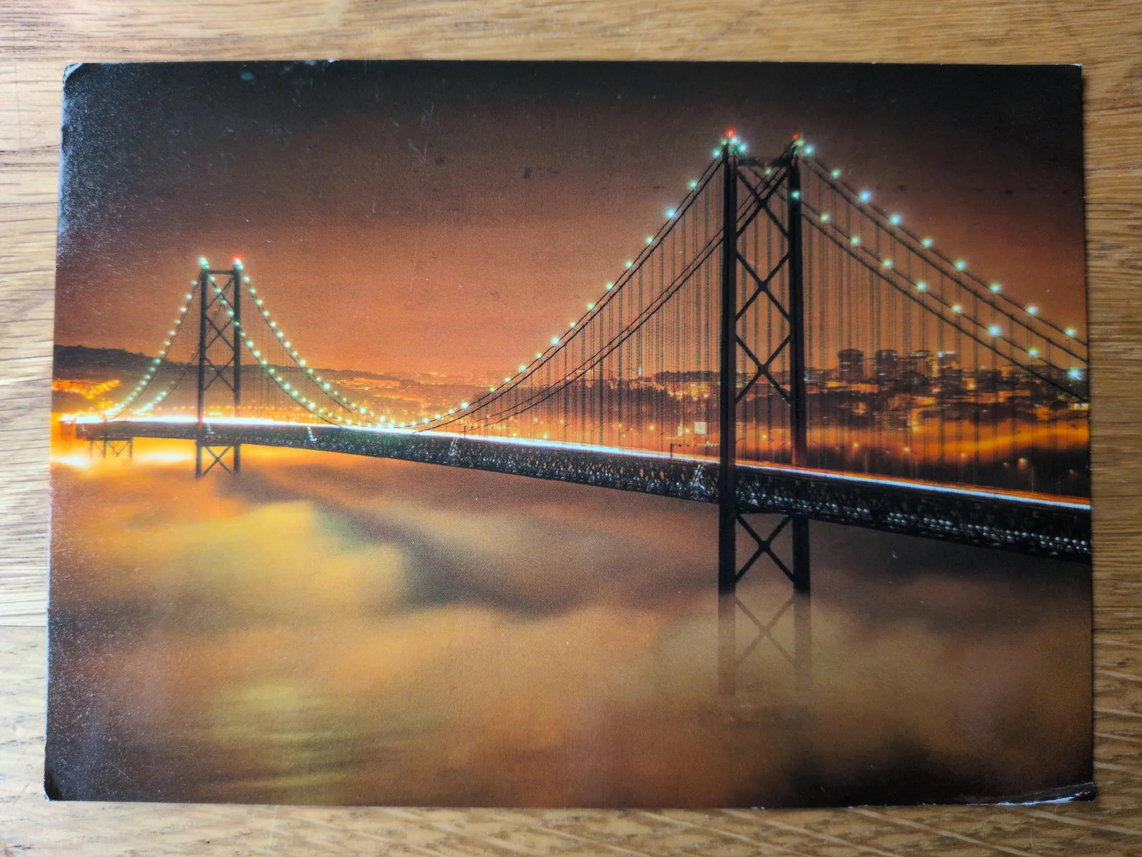 Eine nachts hell erleuchtete Hängebrücke schwebt über orangefarbenem Nebel, mit den Lichtern einer Stadt im Hintergrund.