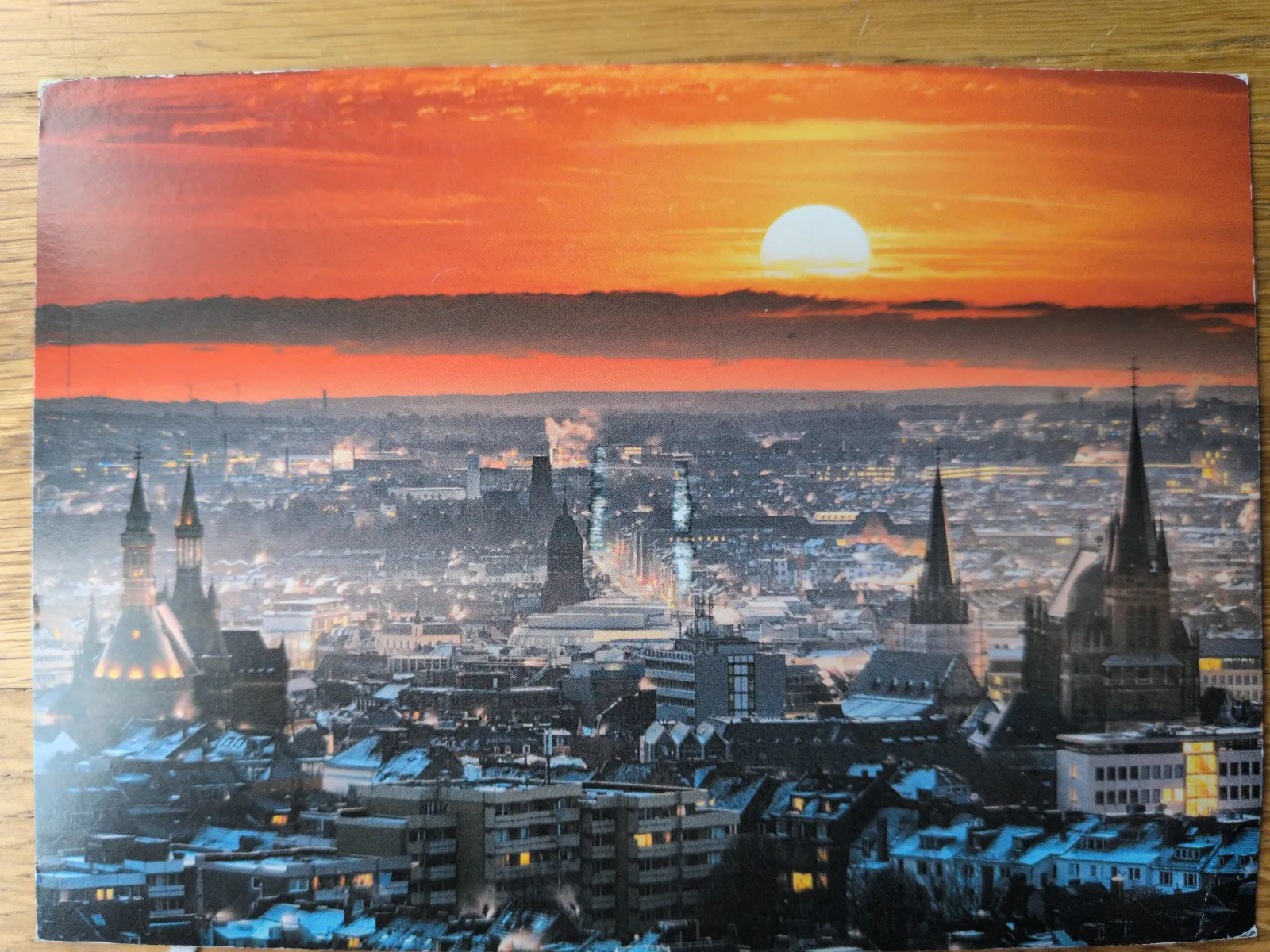 Ein winterliches Stadtpanorama bei Sonnenuntergang mit orange-rotem Himmel, einer hellen Sonne und beleuchteten Gebäuden sowie Kirchtürmen.