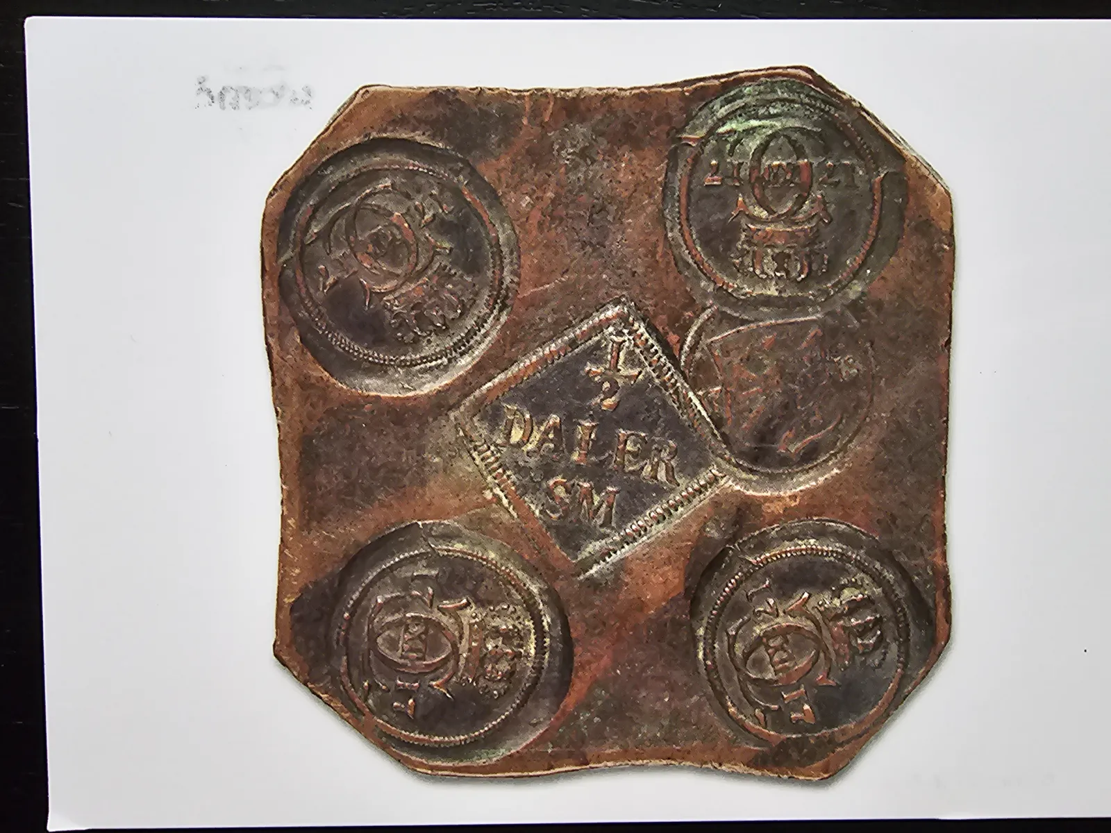 Schwedisches Kupferplatten-Geld (Plåtmynt) von 1717: Eine rechteckige Kupferplatte mit abgeschnittenen Ecken, in jeder Ecke ein geprägter Rundstempel mit dem schwedischen Reichswappen (drei Kronen), in der Mitte ein rautenförmiger Wertstempel mit '1/2 DALER S.M.' (Silvermynt). Die Patina zeigt kupfergrüne und braune Oxidation.