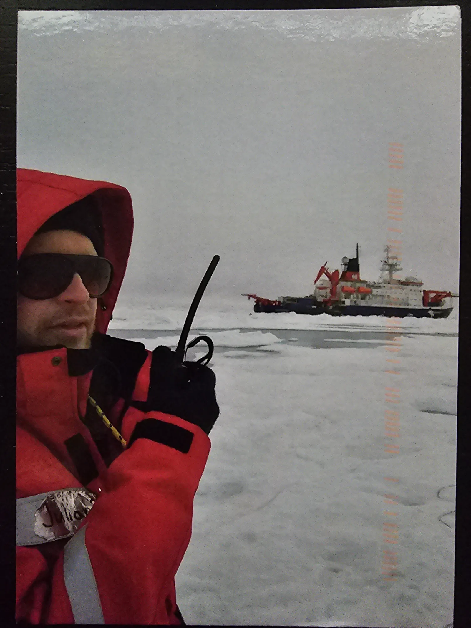 Ein Wissenschaftler in roter Polarexpeditionskleidung mit Kapuze und Sonnenbrille steht auf arktischem Meereis und hält ein Funkgerät. Im Hintergrund liegt der deutsche Forschungseisbrecher FS Polarstern mit seiner markanten rot-weißen Farbgebung. Der Himmel ist bedeckt, die Eislandschaft erstreckt sich bis zum Horizont.