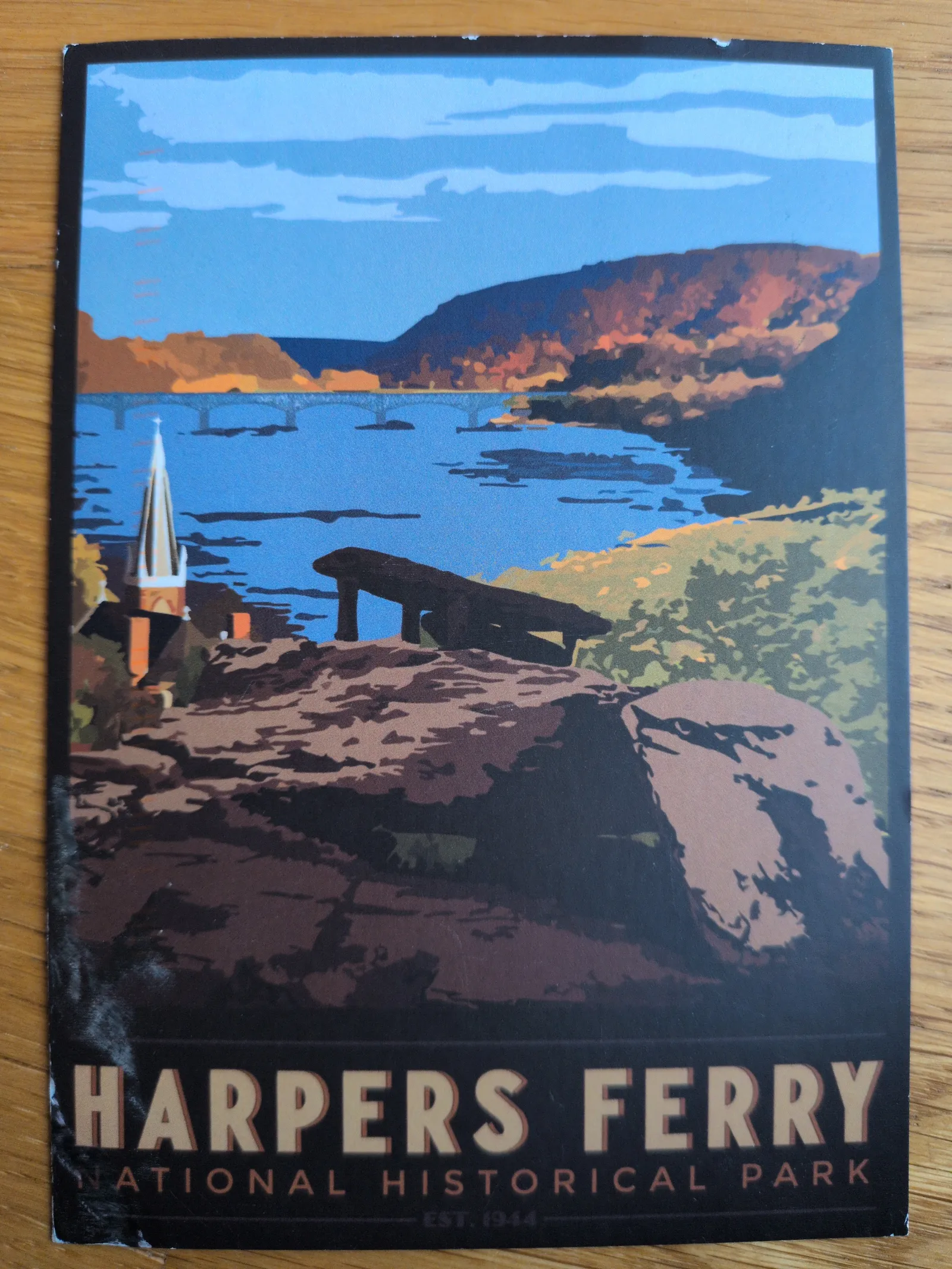 Ein stilisiertes Poster zeigt eine Landschaft von Harpers Ferry mit einem Fluss, einer Brücke, bewaldeten Hügeln, einer Kirche und dem Schriftzug „Harpers Ferry National Historical Park“.