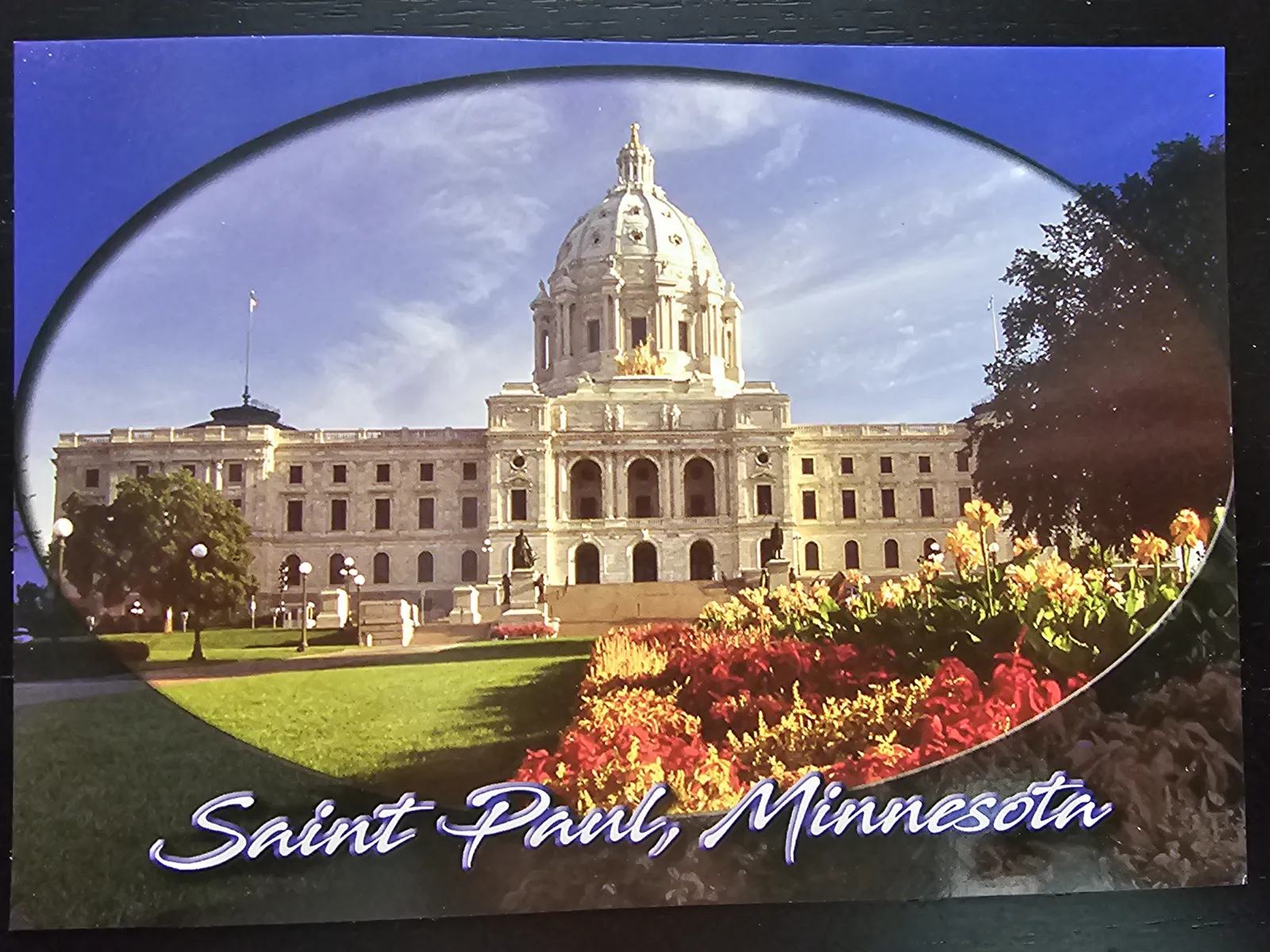 Das imposante Minnesota State Capitol mit seiner großen Kuppel ist von grünen Rasenflächen und bunten Blumenbeeten umgeben, darunter steht "Saint Paul, Minnesota".