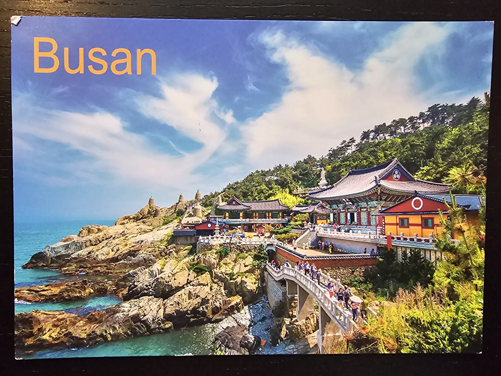 Ein farbenprächtiger koreanischer Tempelkomplex thront auf einer felsigen Küste am blauen Meer unter einem bewölkten Himmel, mit dem Schriftzug „Busan“ links oben.