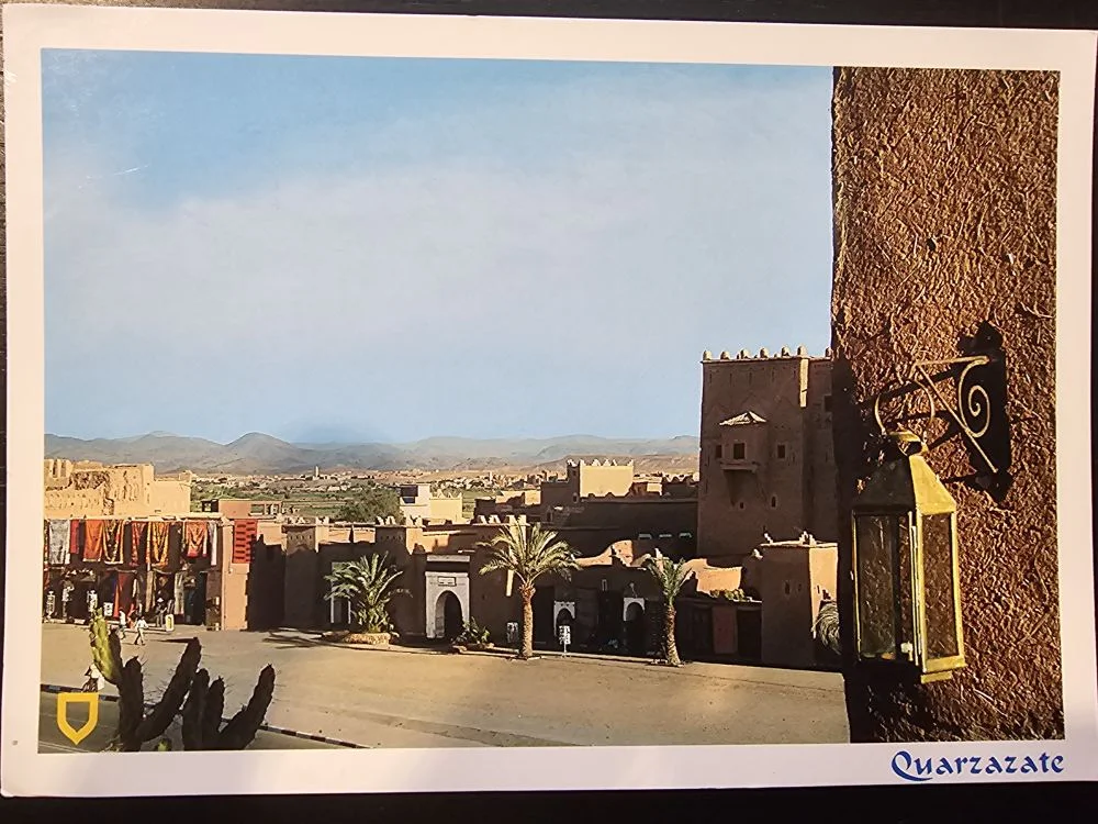 Eine sonnige Ansicht der marokkanischen Stadt Ouarzazate zeigt traditionelle Lehmgebäude, einen weiten Platz mit Palmen, Berge im Hintergrund und eine Laterne rechts im Vordergrund.