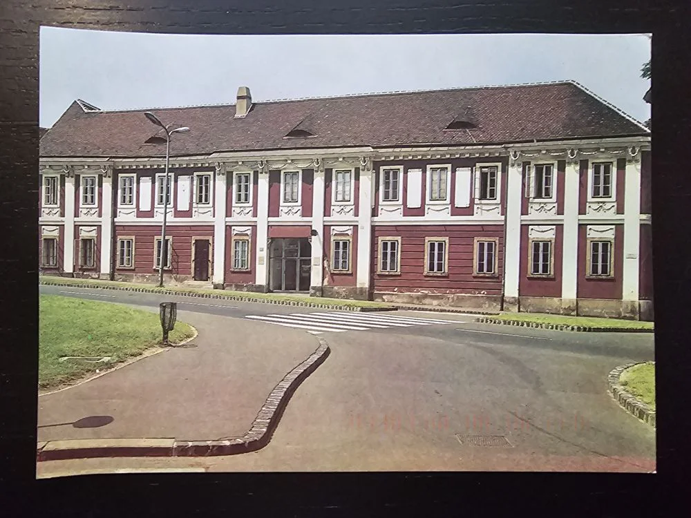 Ein langes, zweistöckiges Gebäude mit rotem Dach, roten Wänden und weißen Fensterrahmen steht entlang einer Straße mit Zebrastreifen und Grünflächen.
