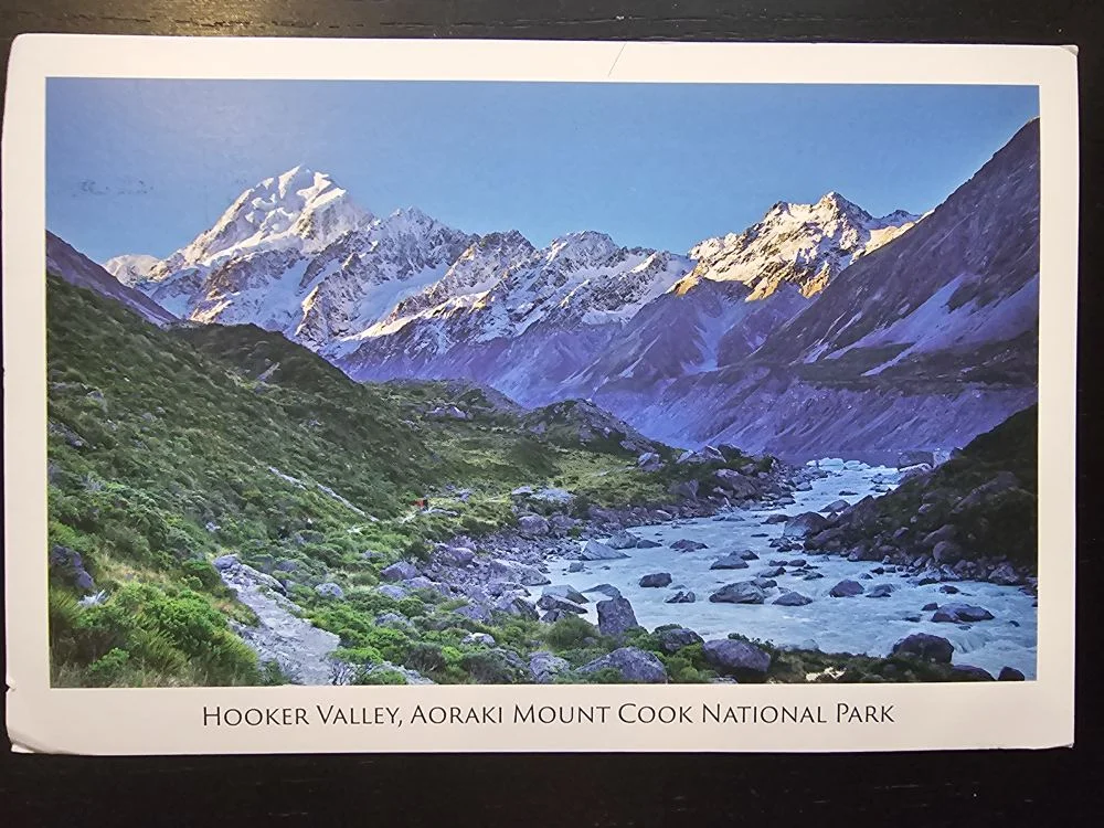 Ein grünes Tal mit einem Fluss und Wanderweg erstreckt sich vor majestätischen, schneebedeckten Bergen unter blauem Himmel.