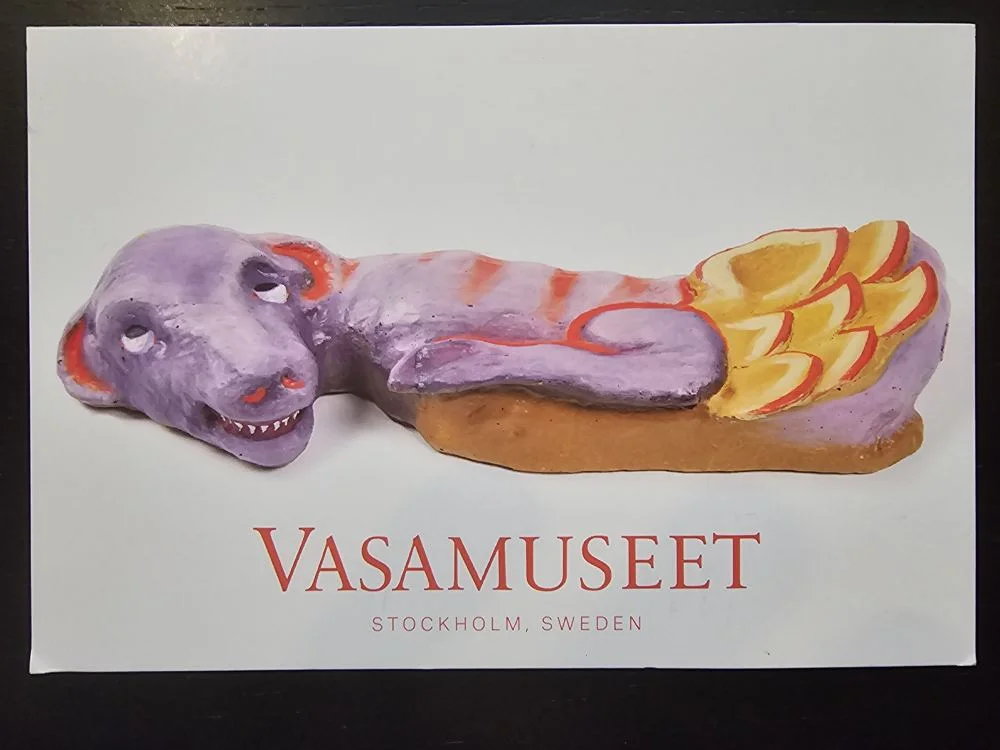 Ein Postkartenbild zeigt eine lila-rote, drachenähnliche Figur mit gelben Flossen und spitzen Zähnen, liegend auf einem braunen Sockel, darunter der Schriftzug „VASAMUSEET STOCKHOLM, SWEDEN“.