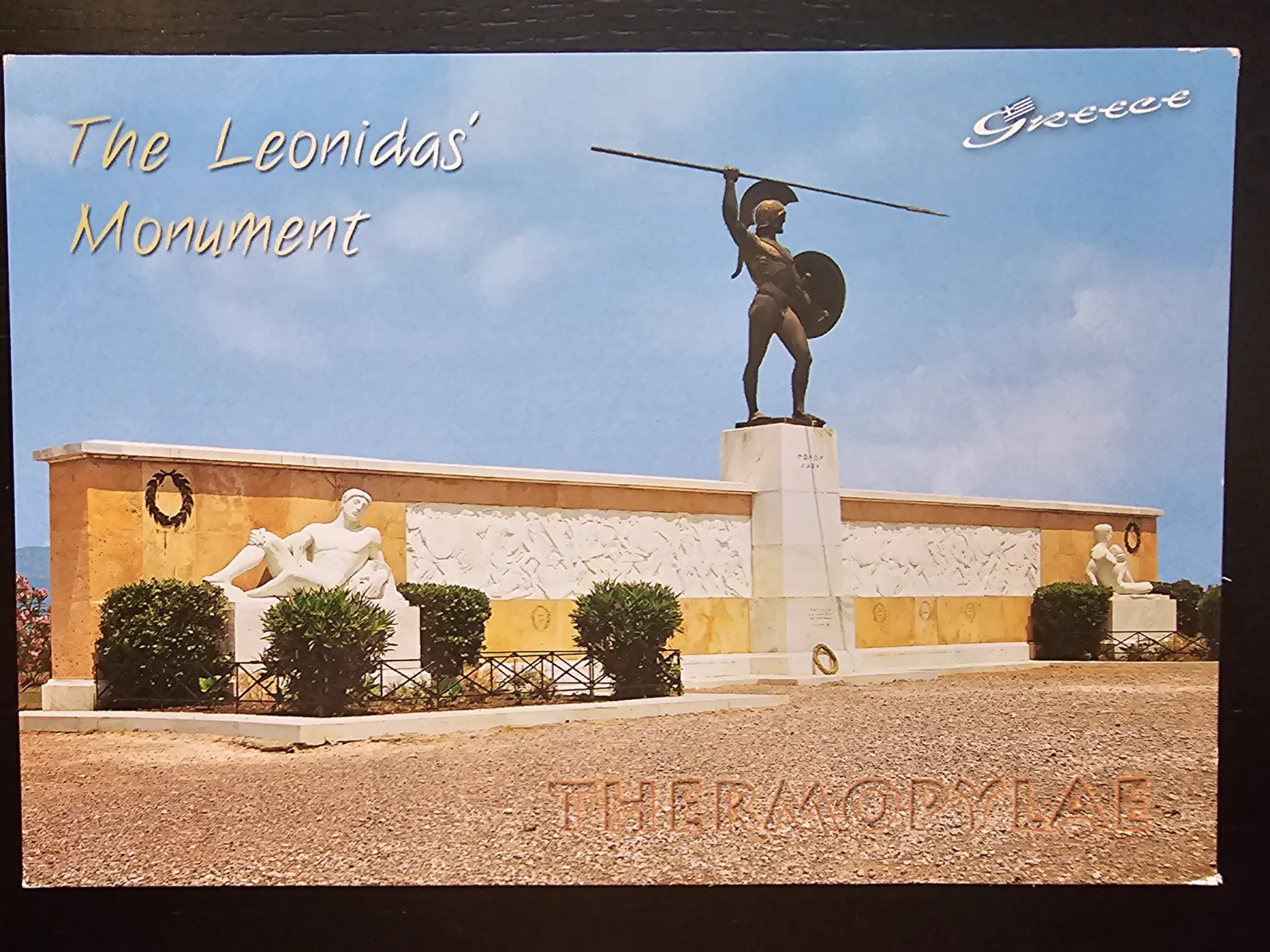 Das Bild zeigt das Leonidas-Denkmal in Thermopylae mit einer großen Bronzestatue eines spartanischen Kriegers, umgeben von weiteren Skulpturen und einer Reliefmauer unter blauem Himmel.