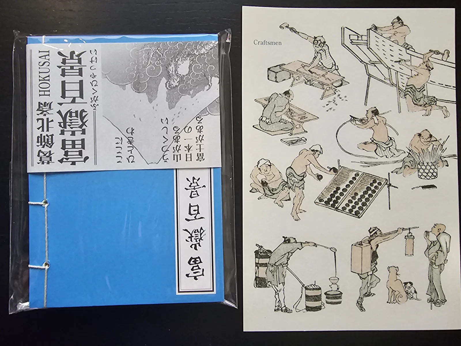 Ein blaues, in Folie verpacktes Büchlein mit japanischer Schrift und dem Namen Hokusai liegt neben einem Blatt mit farbigen Zeichnungen von Handwerkern bei der Arbeit.