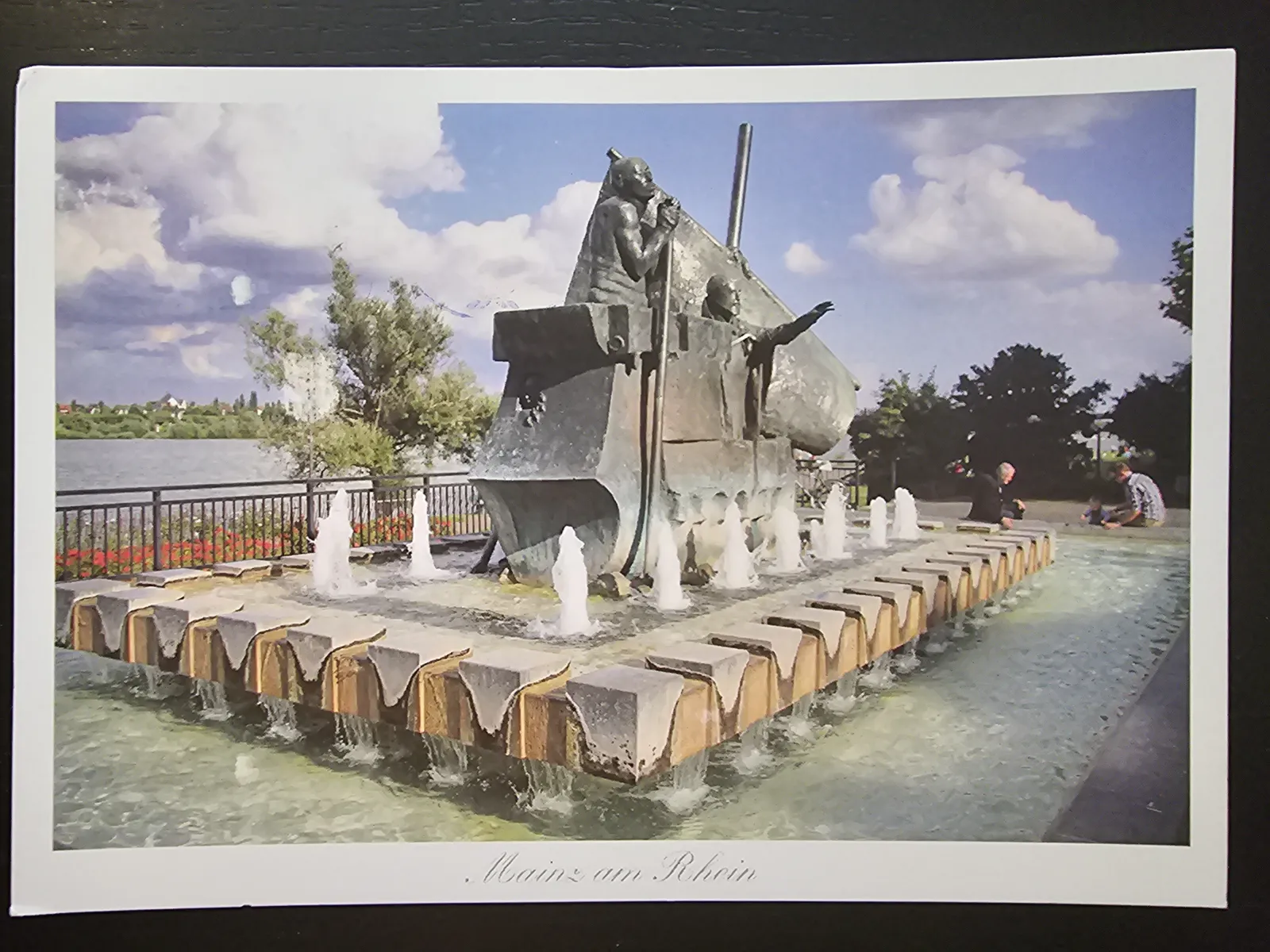 Ein großer, moderner Brunnen in Mainz am Rhein zeigt eine Skulptur von Figuren in einem Schiff, umgeben von vielen Wasserfontänen, Grün und einigen Menschen im Hintergrund.