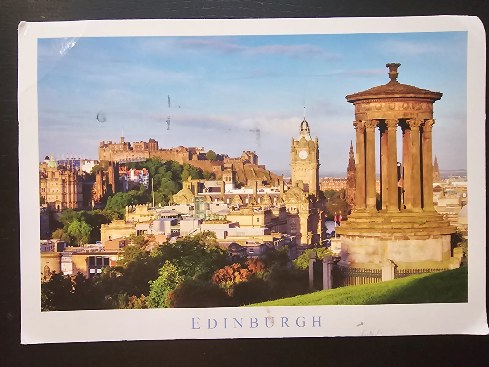 Edinburgh mit einem Säulenmonument auf einem grünen Hügel im Vordergrund, der Stadt mit Schloss und Uhrturm im Hintergrund und dem Schriftzug "EDINBURGH" am unteren Rand.