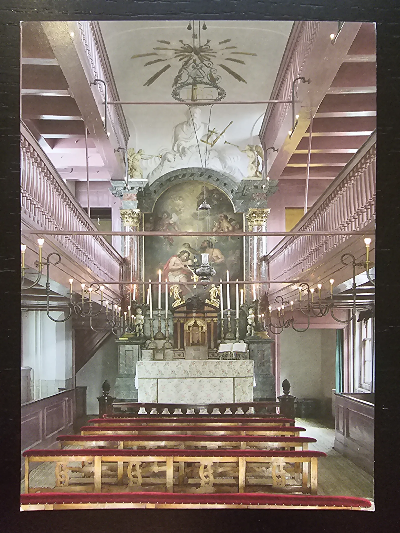 Ein reich verzierter Kirchenraum mit einem Altar, Kerzen, einem großen Gemälde und roten Holzbänken, flankiert von Emporen.