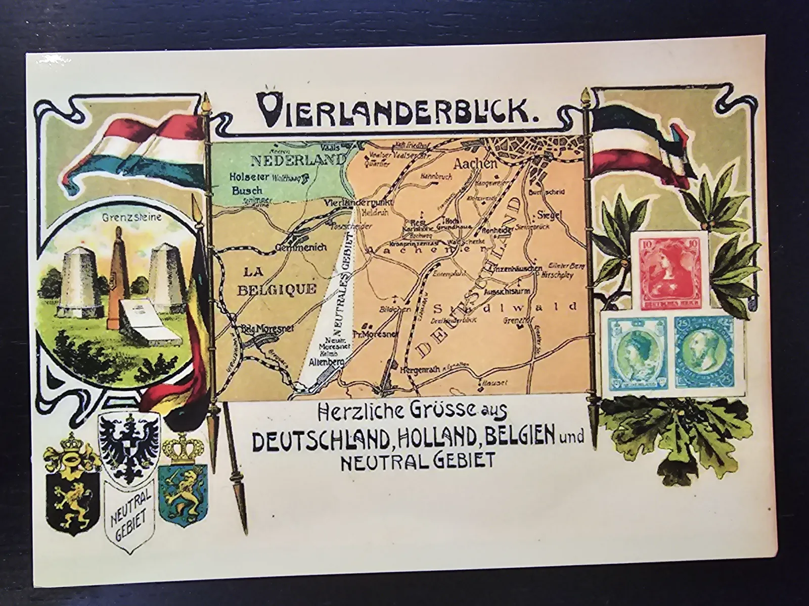 Eine historische Postkarte zeigt eine Karte des Vierländergebiets um Aachen, mit Deutschland, Holland, Belgien und dem Neutralen Gebiet, umrahmt von ihren Flaggen, Wappen und Briefmarken.