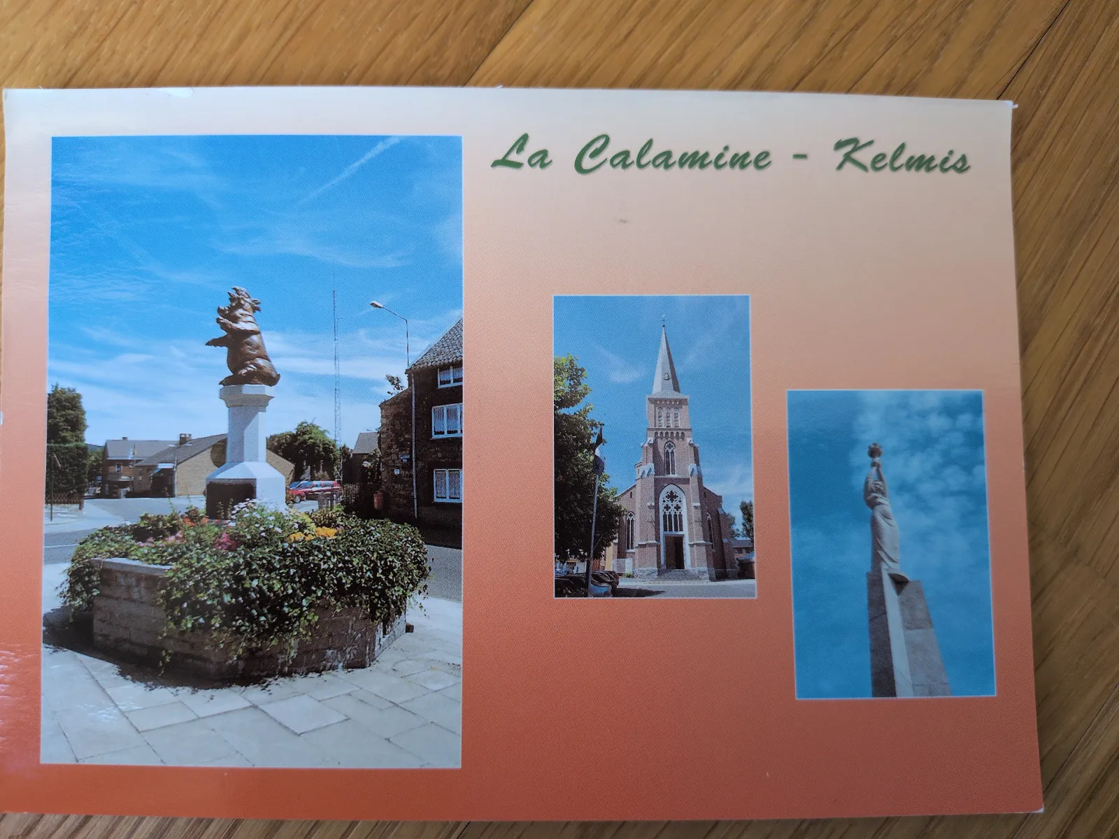 Eine Postkarte zeigt drei Ansichten von Kelmis/La Calamine: eine Schweinestatue auf einem Sockel, eine Kirche mit spitzem Turm und ein hohes Denkmal vor blauem Himmel.