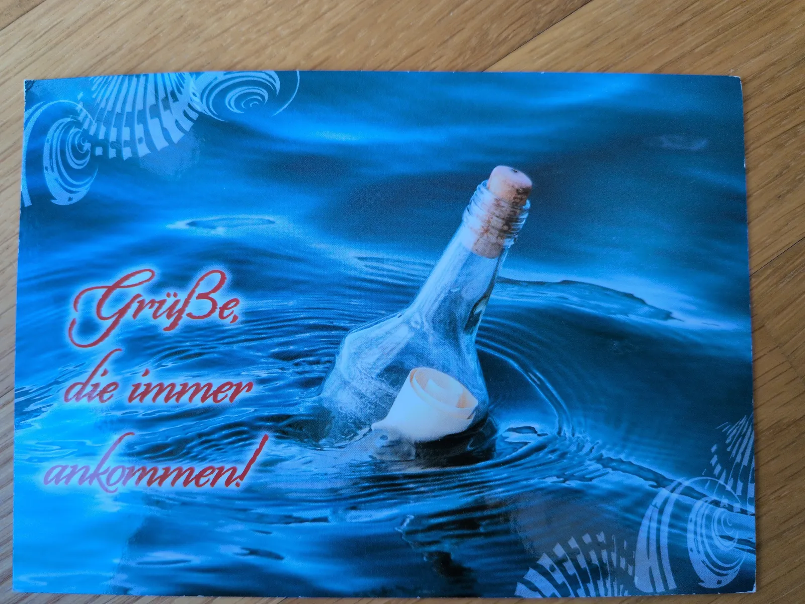 Das Bild zeigt eine Flaschenpost in dunklem, welligem Blauwasser mit dem roten Schriftzug "Grüße, die immer ankommen!".