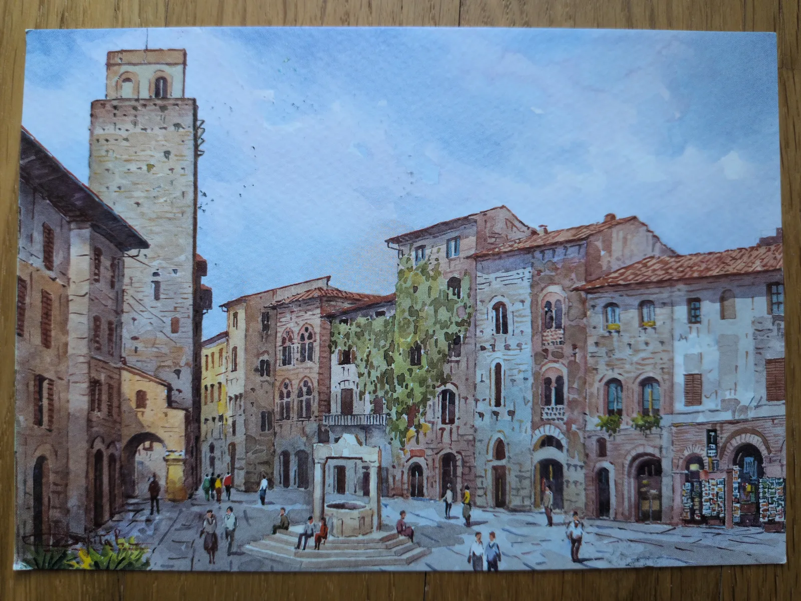 Ein Aquarellgemälde zeigt einen historischen Stadtplatz in San Gimignano mit einem hohen Turm, alten Gebäuden, einem Brunnen und mehreren Personen unter blauem Himmel.