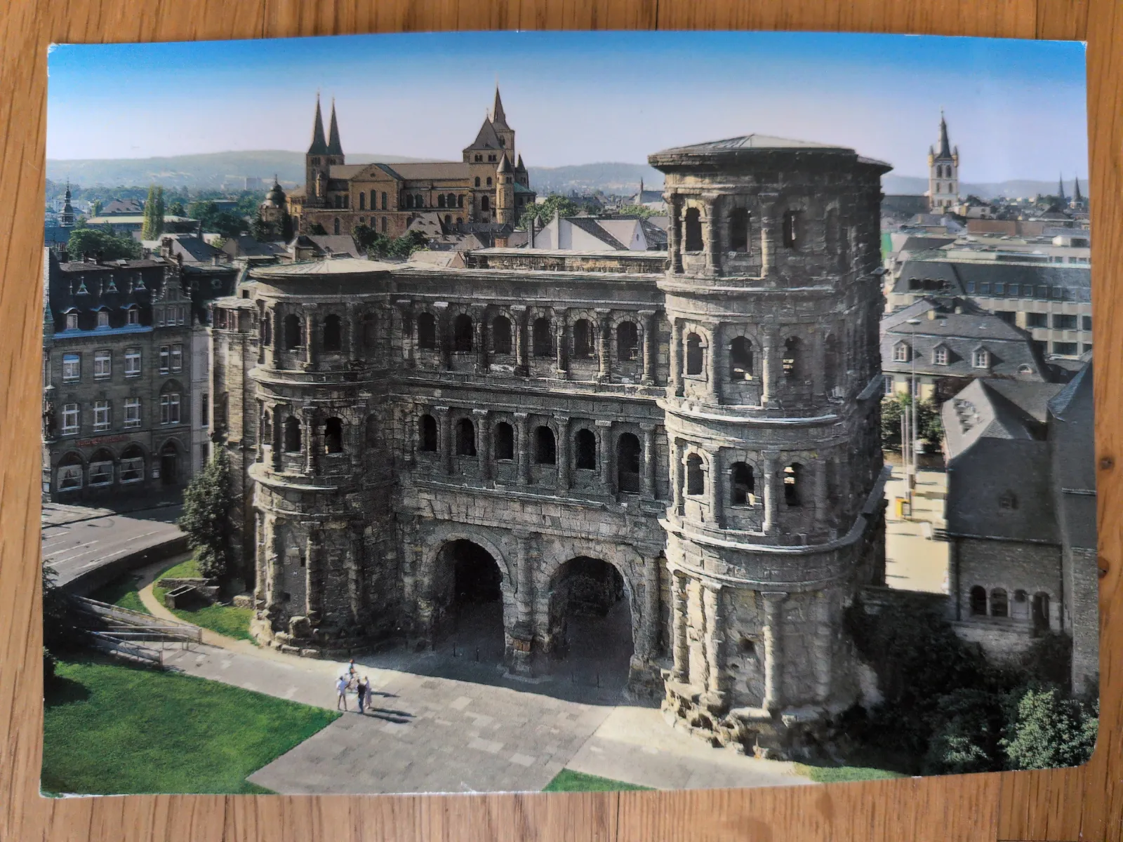 Die Porta Nigra, ein massives römisches Sandsteintor mit zwei Türmen und Bögen, steht im Zentrum des Bildes, dahinter eine Stadtlandschaft mit Kathedrale unter blauem Himmel.