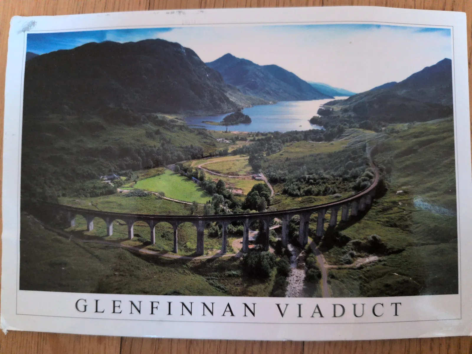 Ein Luftbild zeigt das geschwungene Glenfinnan Viadukt mit seinen Steinbögen in einer weiten grünen Berglandschaft mit einem See im Hintergrund.