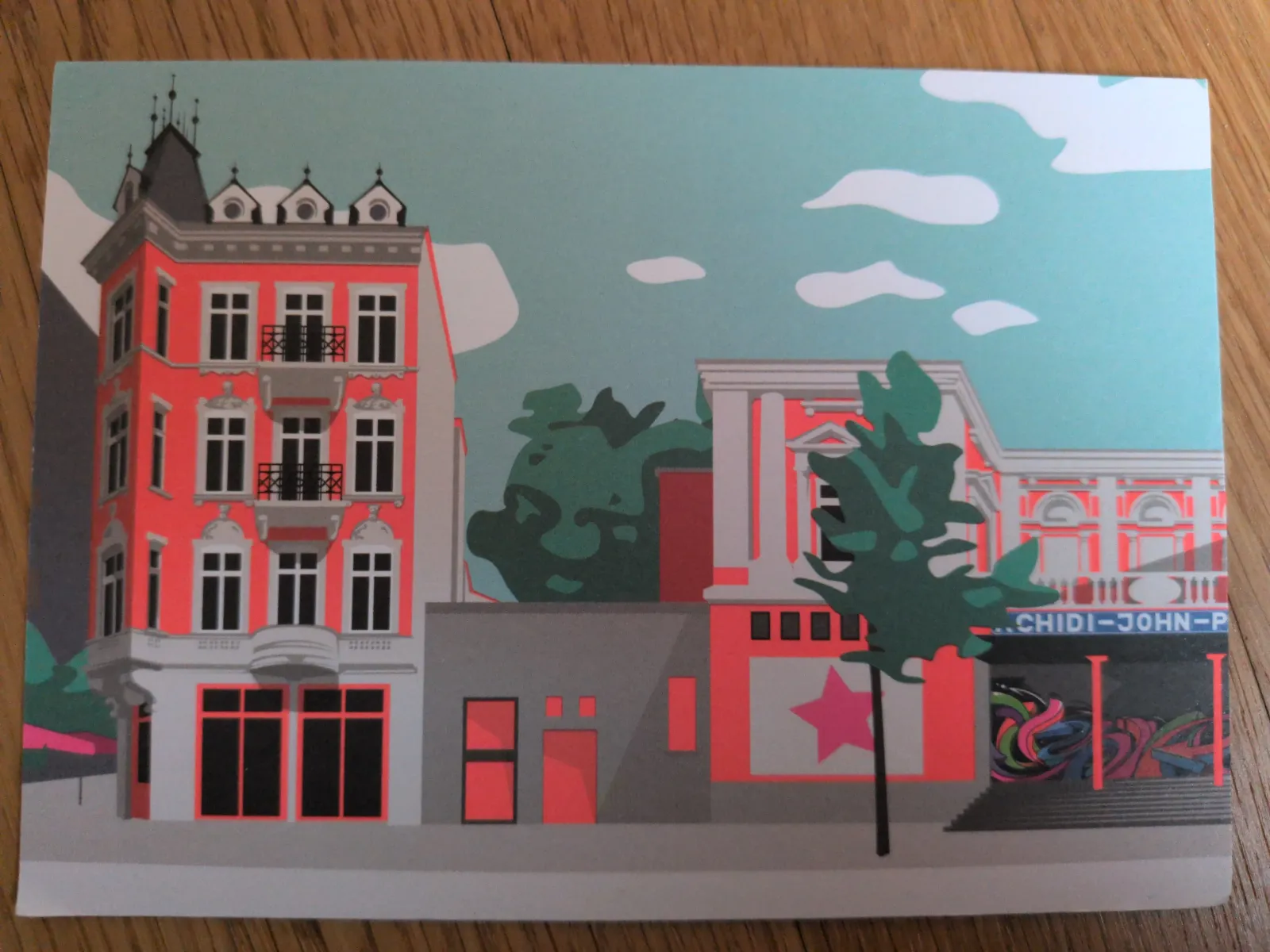 Das Bild zeigt eine stilisierte Stadtansicht mit markanten korallenroten Gebäuden, Bäumen und einem hellblauen Himmel mit Wolken.