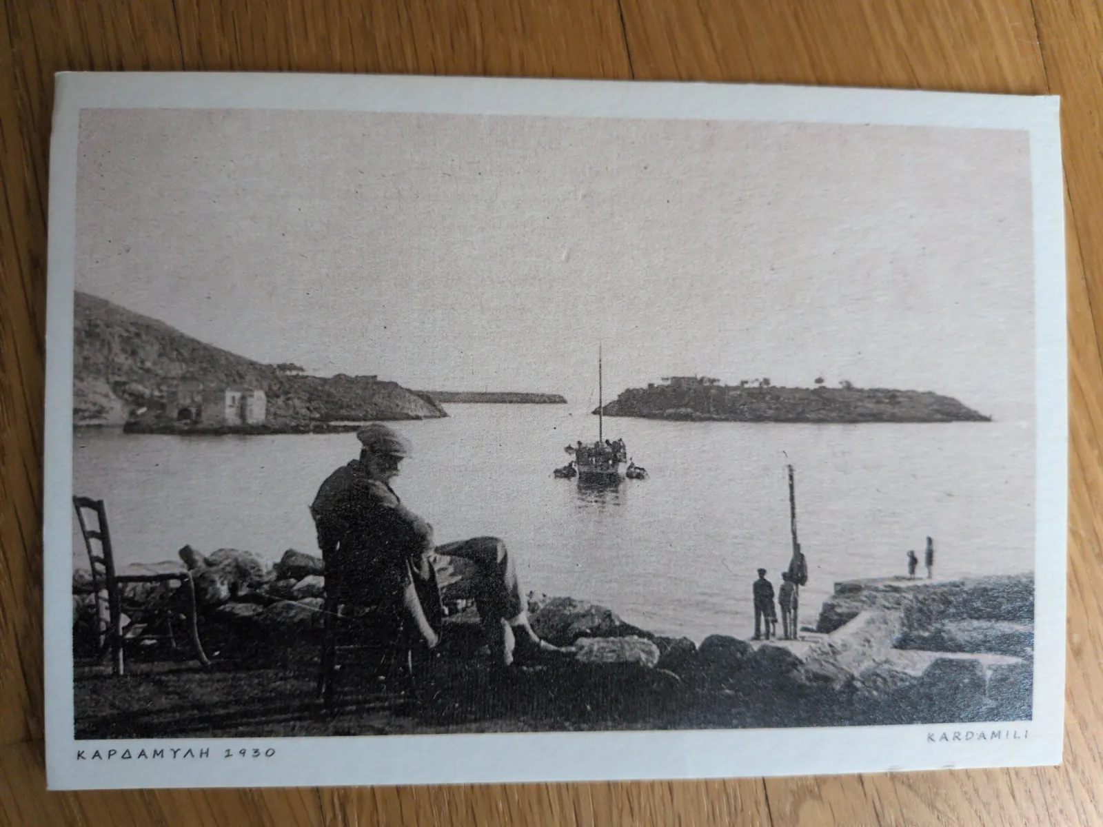 Schwarz-Weiß-Foto einer Bucht in Kardamyli (1930) mit einer sitzenden Person am Ufer, einem Boot auf dem Wasser und einer Insel im Hintergrund.