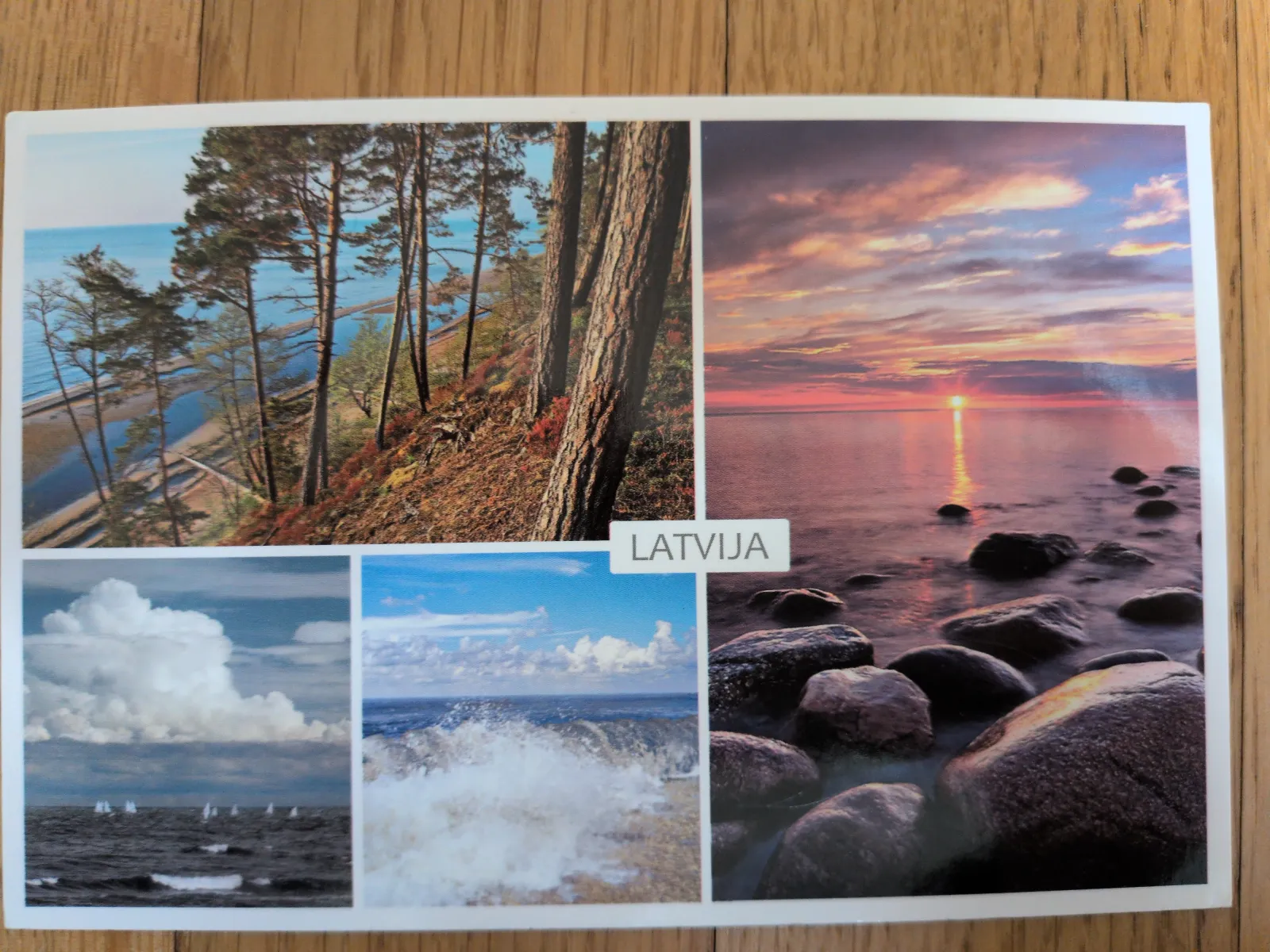 Eine Postkarte zeigt eine Collage von Küstenlandschaften Lettlands mit Kiefernwald am Meer, einem Sonnenuntergang über Felsen, Segelbooten und einer brechenden Welle.