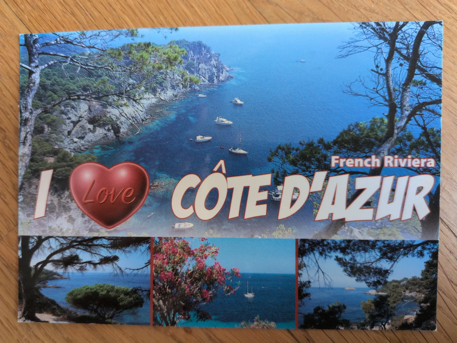 Eine Postkarte mit der Aufschrift „I Love CÔTE D'AZUR“ (wobei „Love“ durch ein rotes Herz ersetzt ist) und „French Riviera“ zeigt eine malerische Felsenküste mit blauem Meer, Booten und Bäumen.