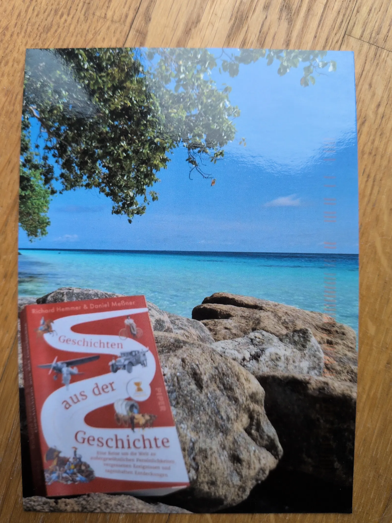 Ein rotes Buch mit dem Titel „Geschichten aus der Geschichte“ liegt auf Felsen an einem tropischen Strand mit blauem Meer und Himmel.