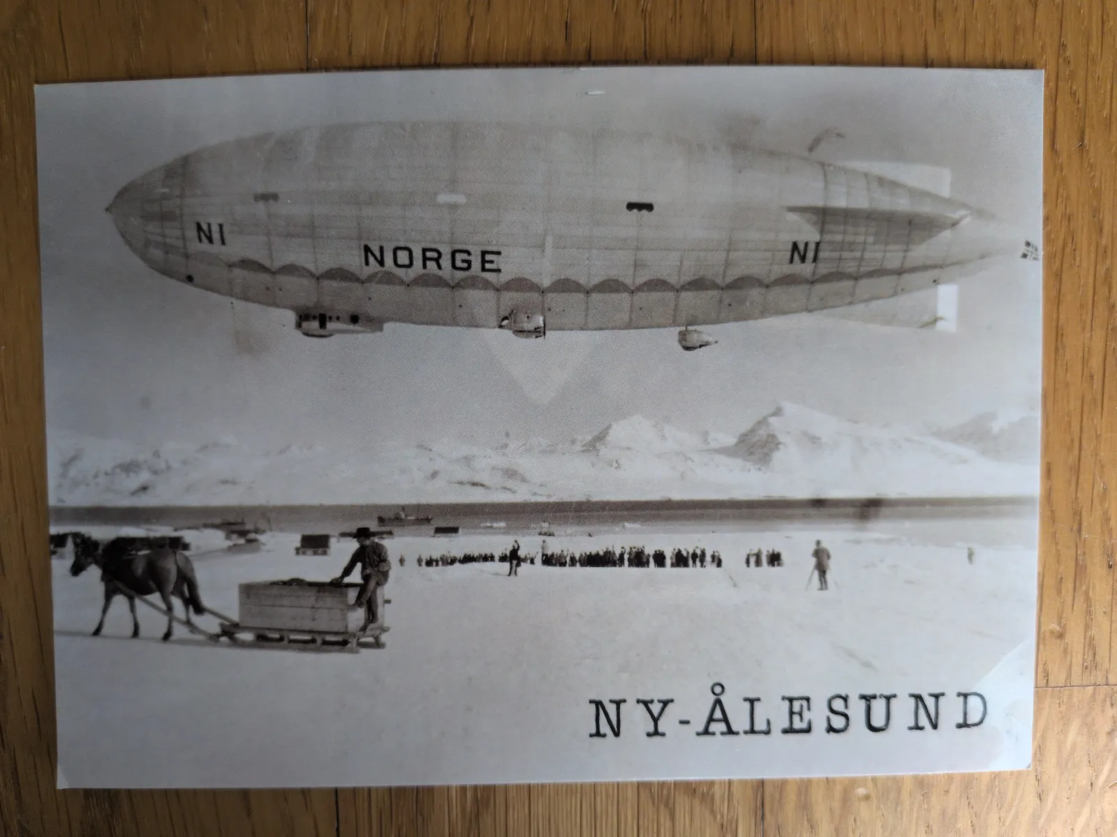 Ein Schwarz-Weiß-Foto zeigt das Luftschiff NORGE über dem verschneiten Ny-Ålesund, mit Menschen und einem Pferdeschlitten auf dem Eis.