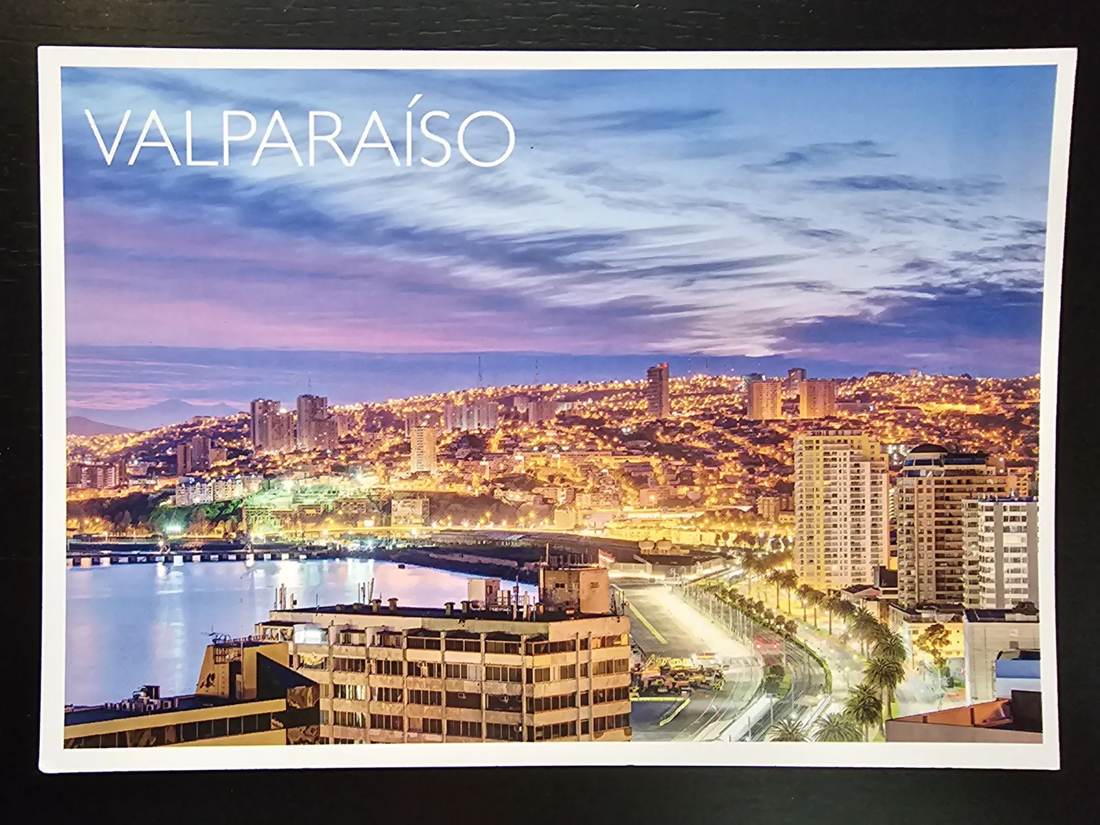Ein nächtliches Panorama von Valparaíso zeigt die Stadt mit tausenden Lichtern an den Hügeln, einem ruhigen Meer und einem farbenfrohen Abendhimmel.