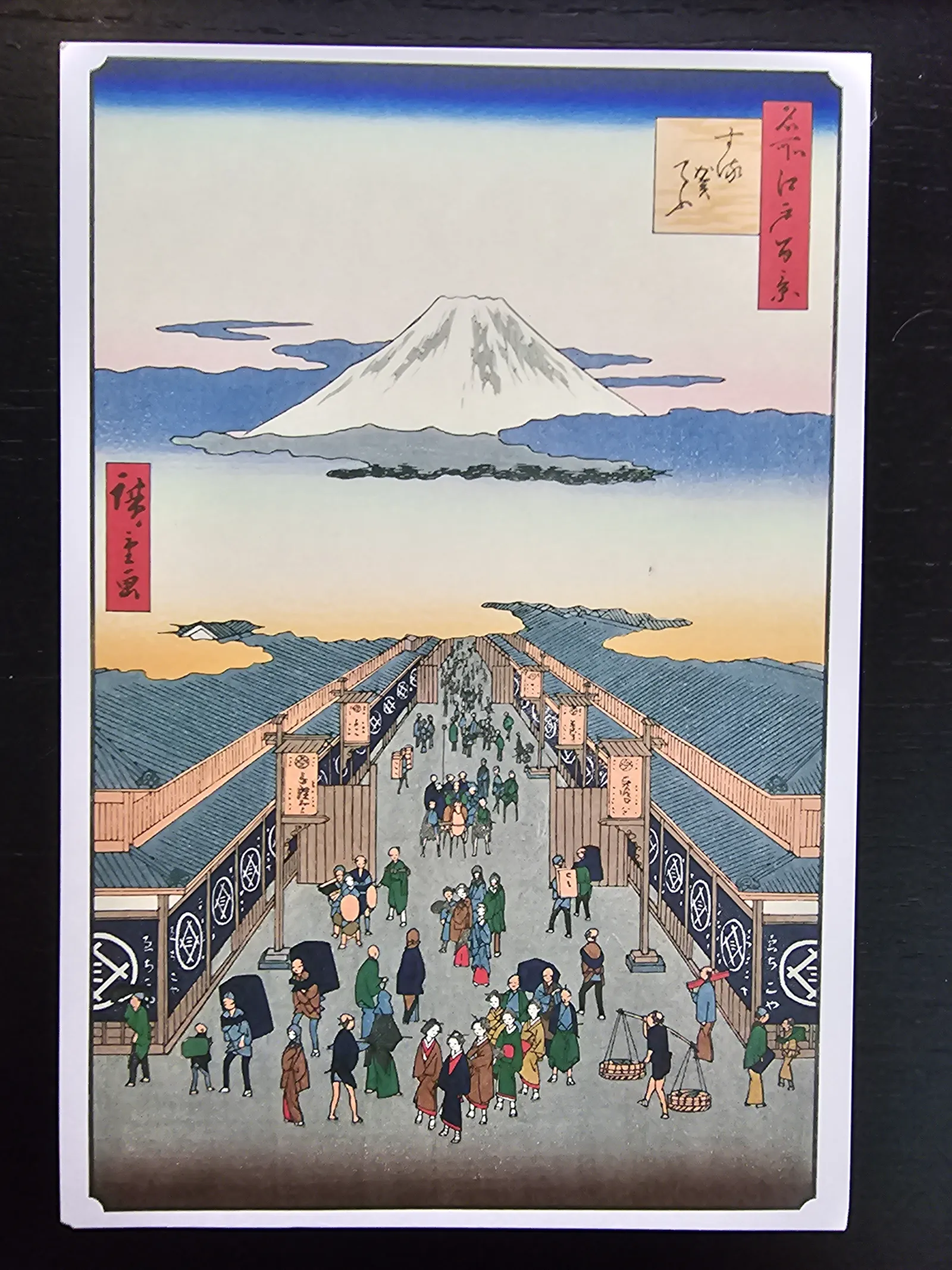 Ein japanischer Holzschnitt zeigt eine belebte Straße mit traditionellen Gebäuden und vielen Menschen, überragt vom schneebedeckten Berg Fuji.