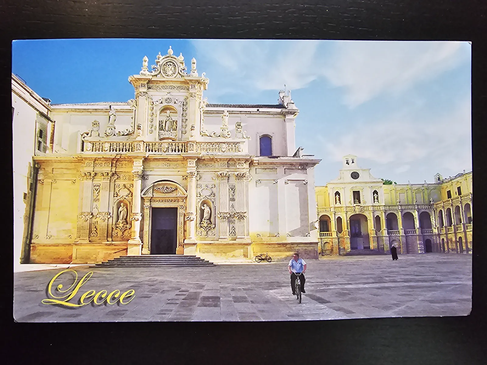 Ein sonniger Platz in Lecce mit einer prunkvollen Barockkirche, weiteren Gebäuden und einem Radfahrer.