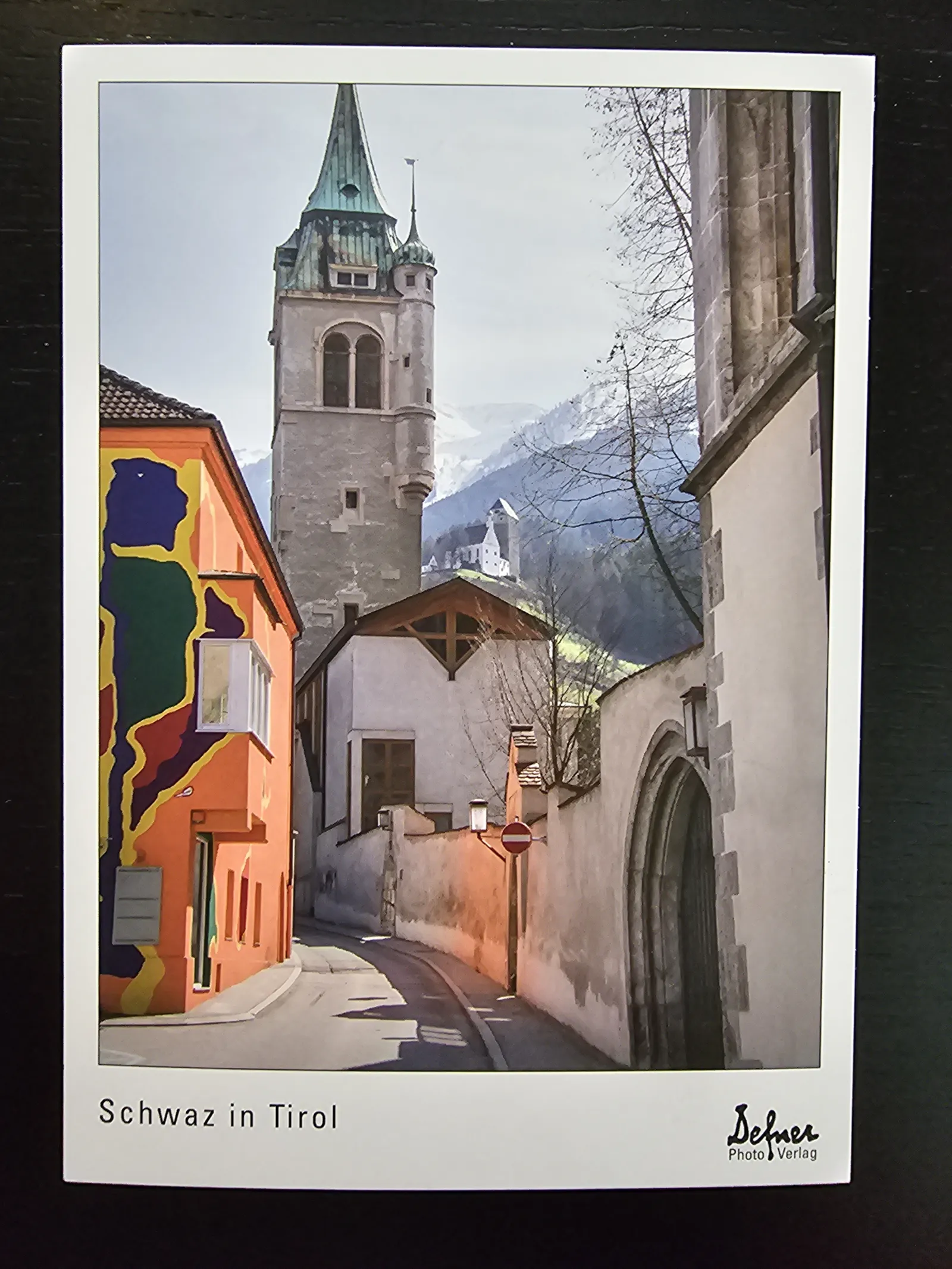 Eine malerische Gasse in Schwaz, Tirol, mit einem hohen Kirchturm, einem farbenfroh bemalten Haus links und schneebedeckten Bergen im Hintergrund.