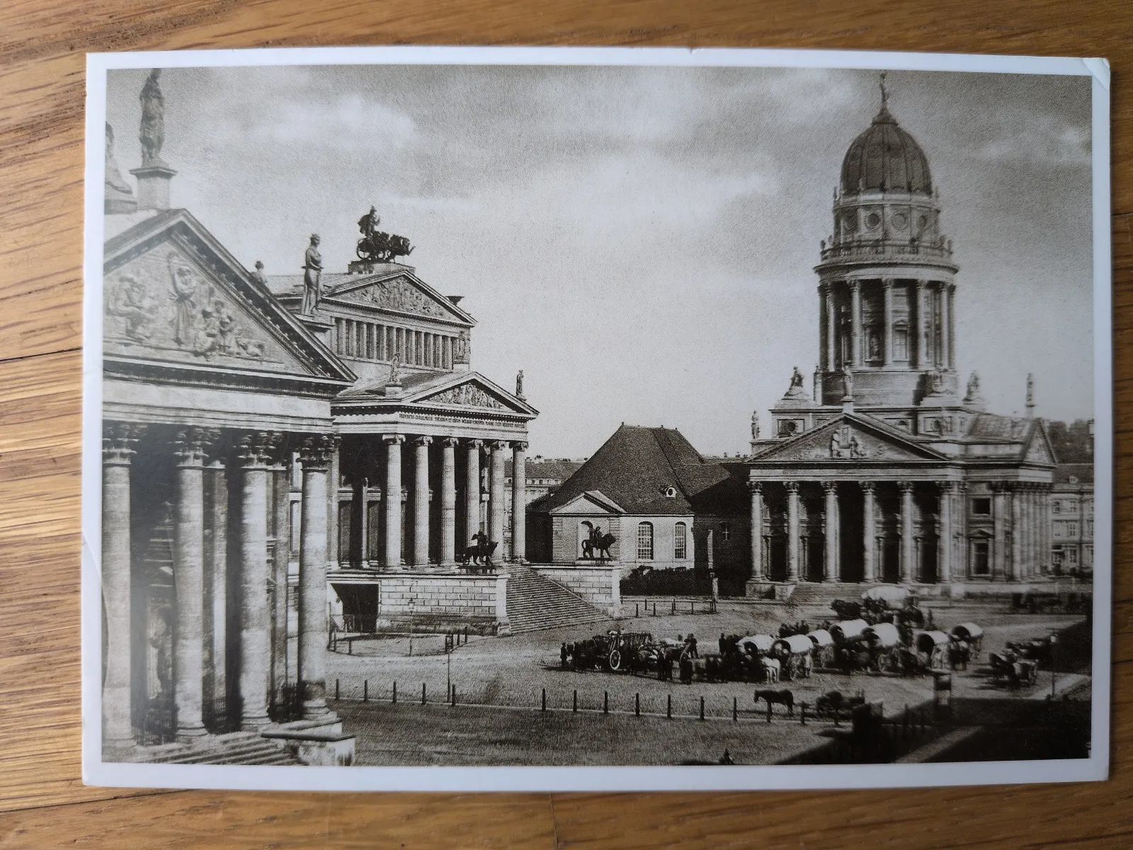 Ein historisches Schwarz-Weiß-Foto zeigt einen großen Platz mit imposanten klassizistischen Gebäuden, darunter eines mit einer Kuppel, sowie Pferdewagen und Menschen.