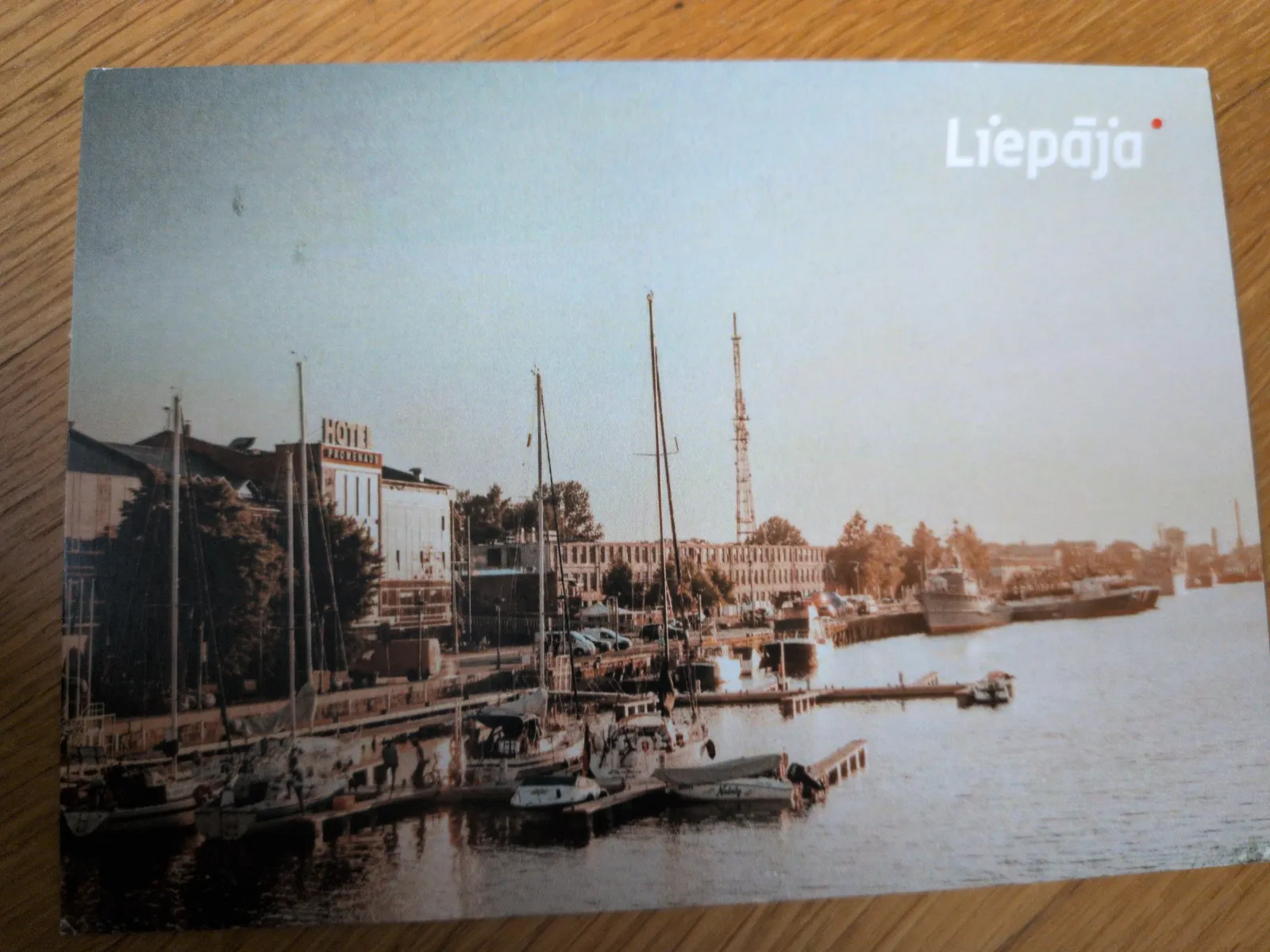 Das Bild zeigt eine Hafenansicht von Liepāja mit zahlreichen Segelbooten, Gebäuden am Ufer und einem hohen Sendeturm unter hellem Himmel.