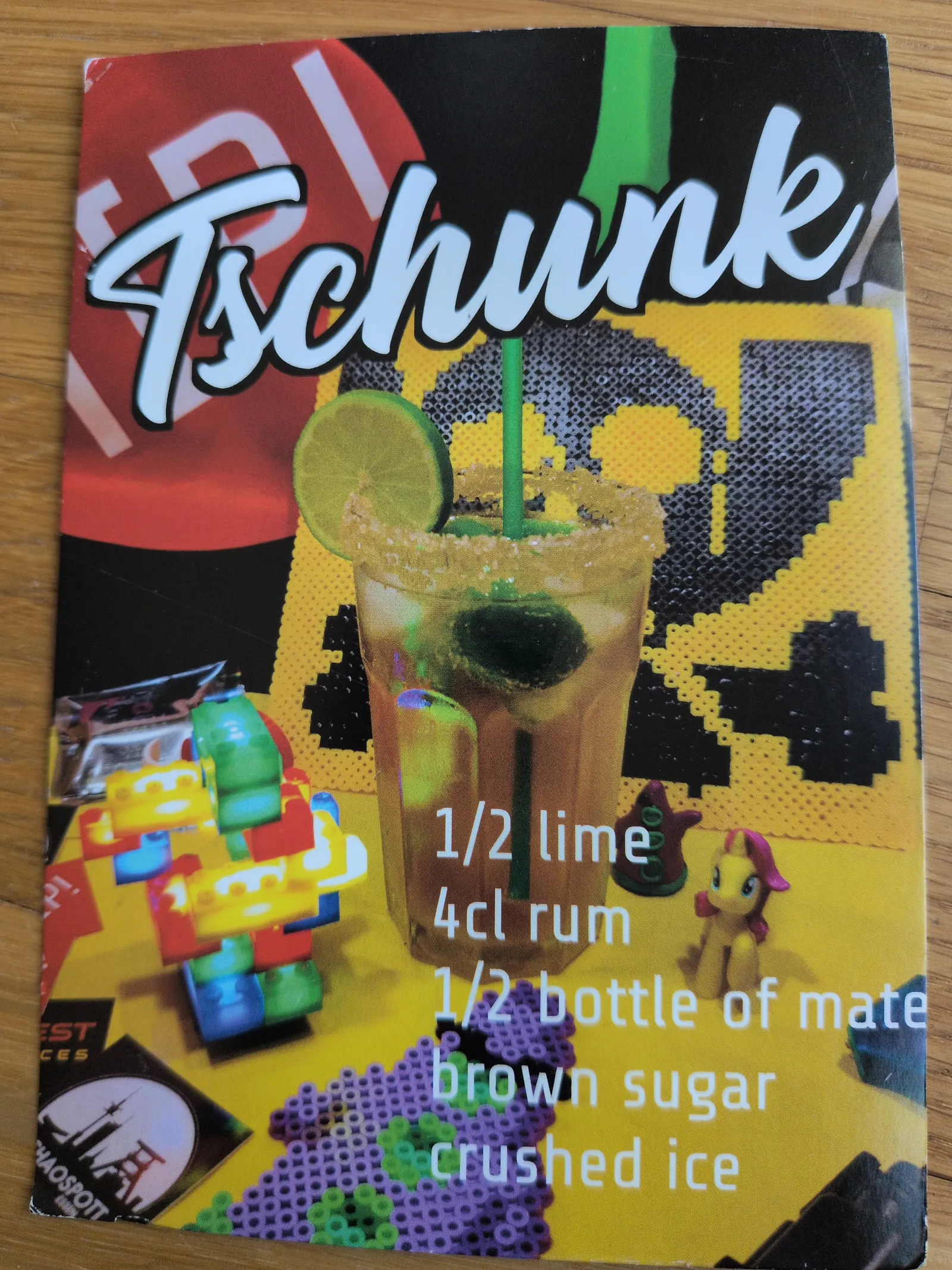 Ein Cocktailrezept für "Tschunk" mit Limette, Rum und Mate ist auf einem bunten Hintergrund mit Spielzeugfiguren und einem pixeligen Totenkopf dargestellt.