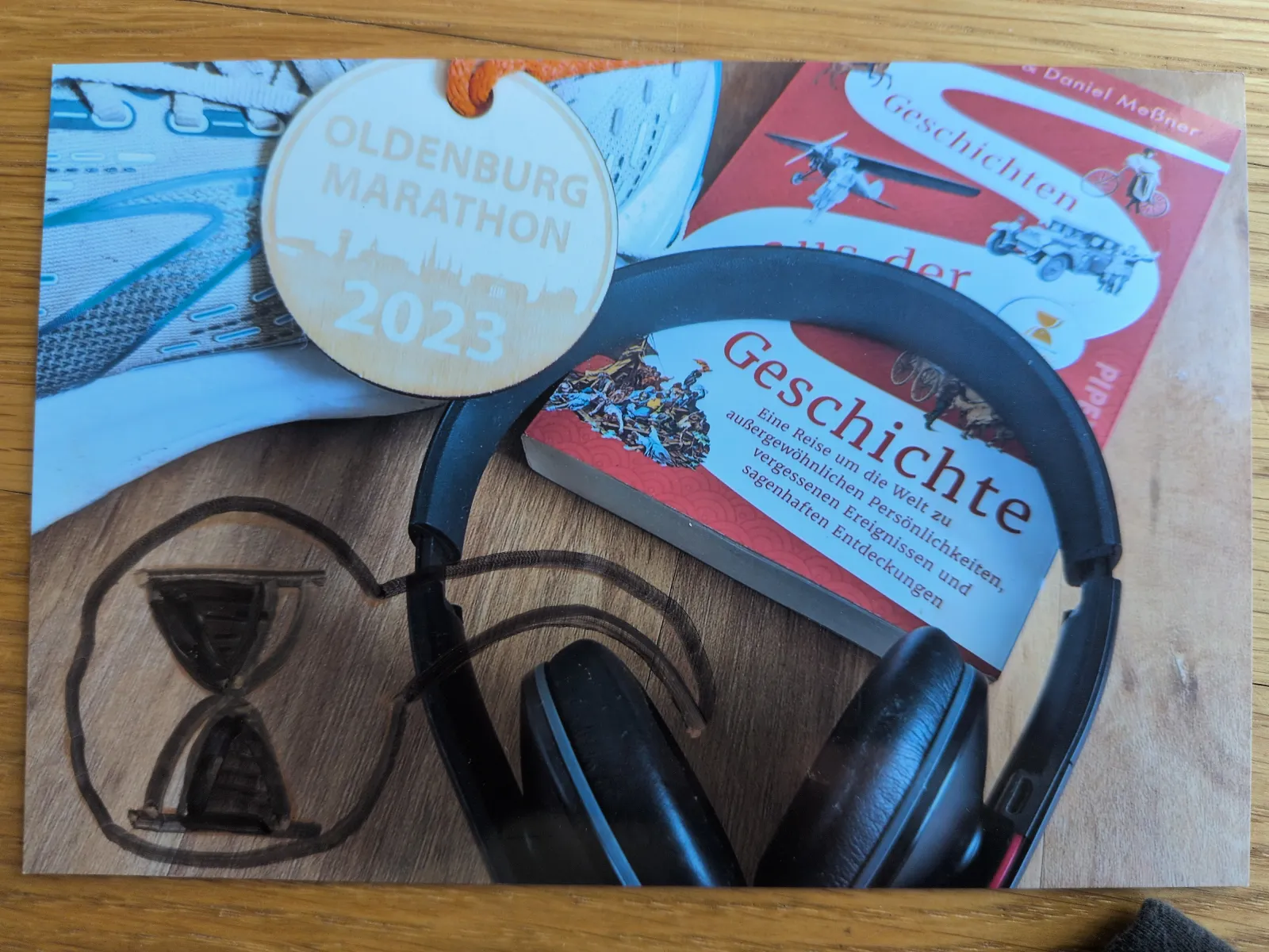 Auf einem Holzhintergrund liegen ein Laufschuh, eine Oldenburg Marathon 2023 Medaille, ein Buch mit dem Titel "Geschichte", schwarze Kopfhörer und ein gezeichnetes Sanduhr-Symbol.