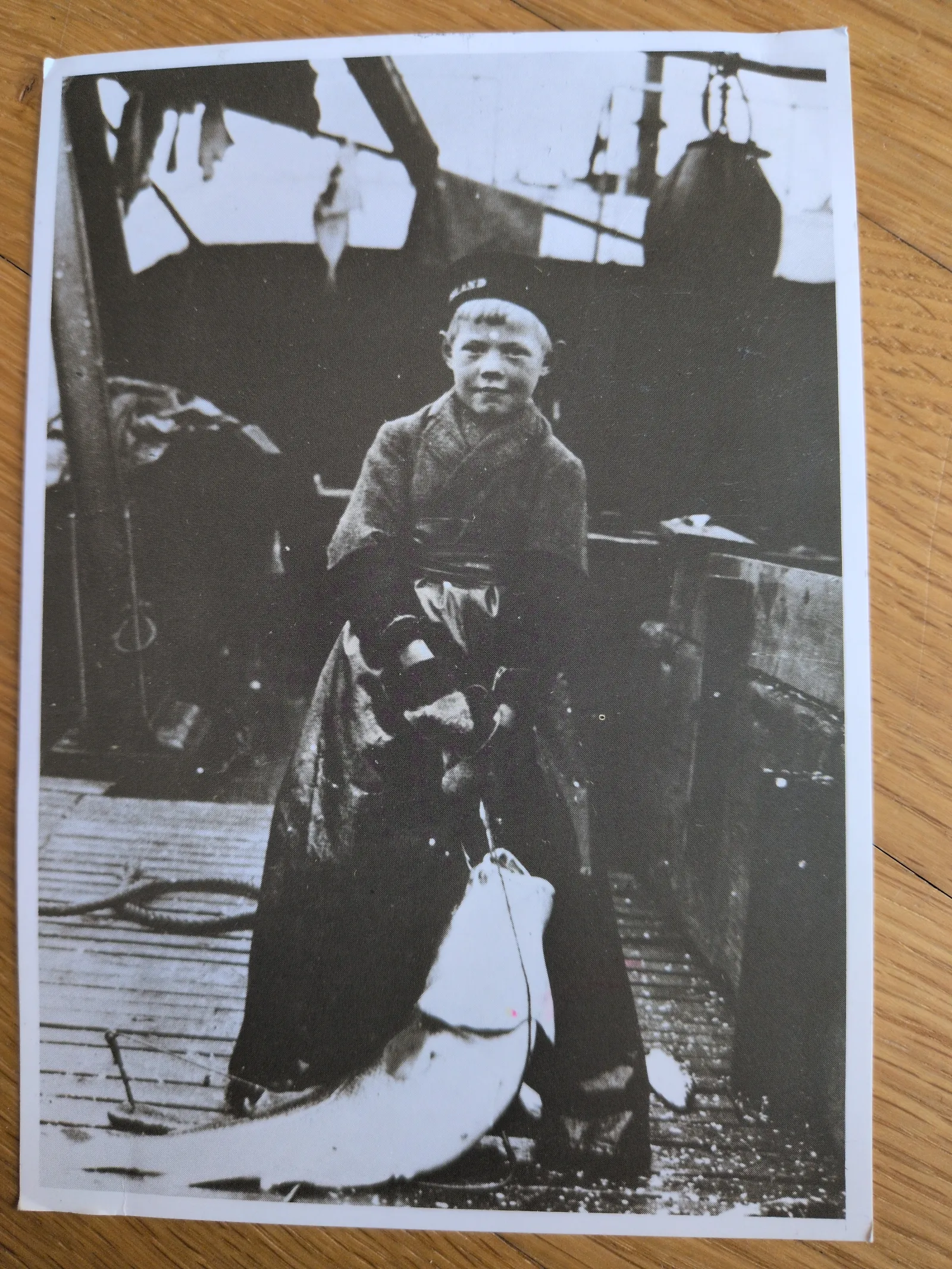 Ein Schwarz-Weiß-Bild zeigt einen kleinen Jungen auf einem Holzdeck, der stolz einen riesigen Fisch hält, der fast so groß ist wie er selbst.