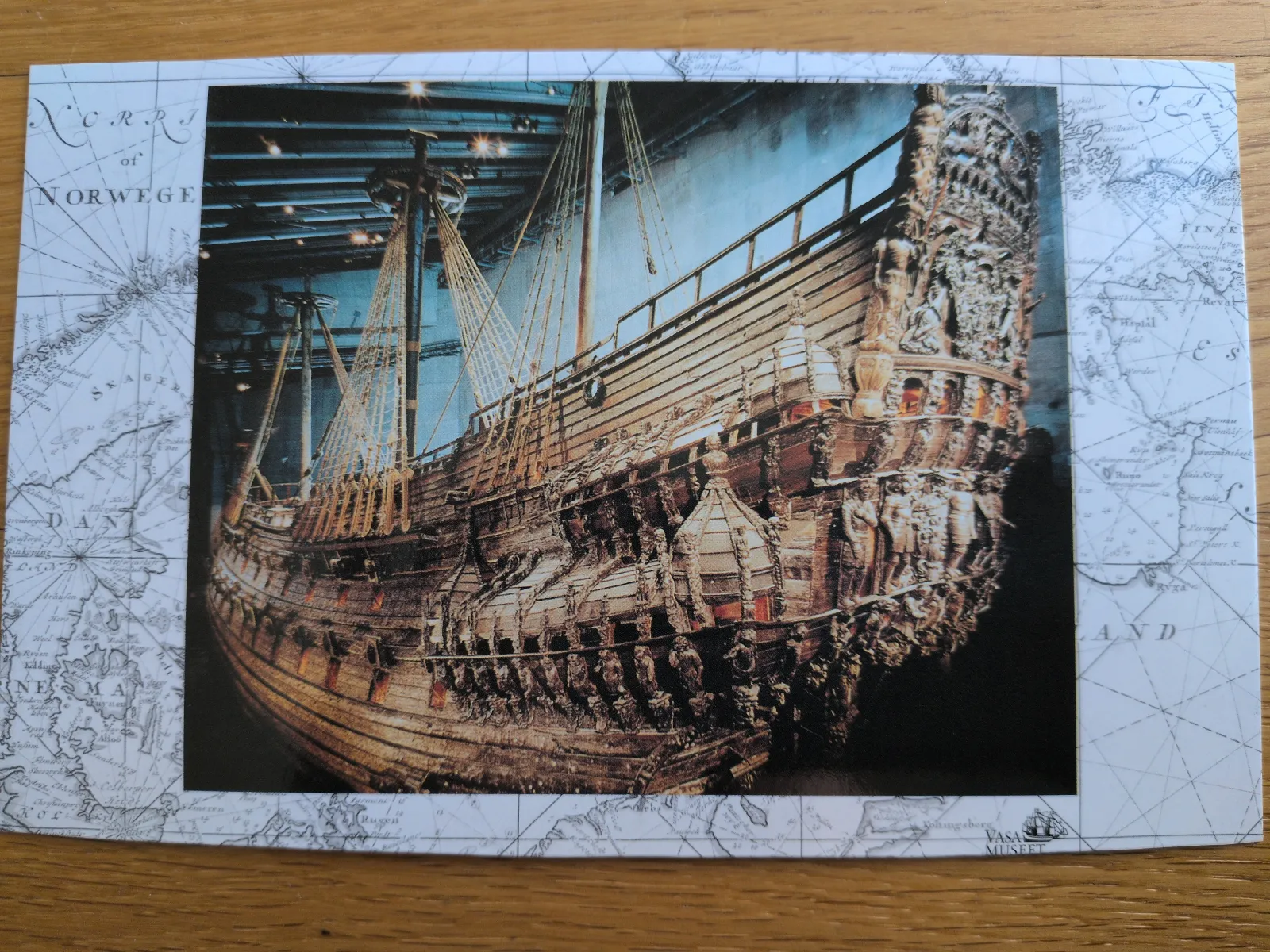 Das Bild zeigt das große, reich verzierte Holzschiff Vasa in einem Museum, umgeben von einer alten Seekarte.