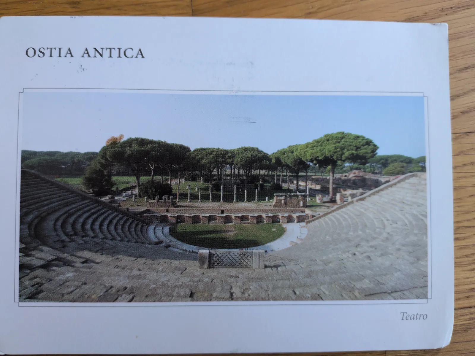 Das Bild zeigt das antike Theater von Ostia Antica mit seinen steinernen Sitzreihen, einer grünen Bühne und dahinterliegenden Ruinen, umgeben von Bäumen.