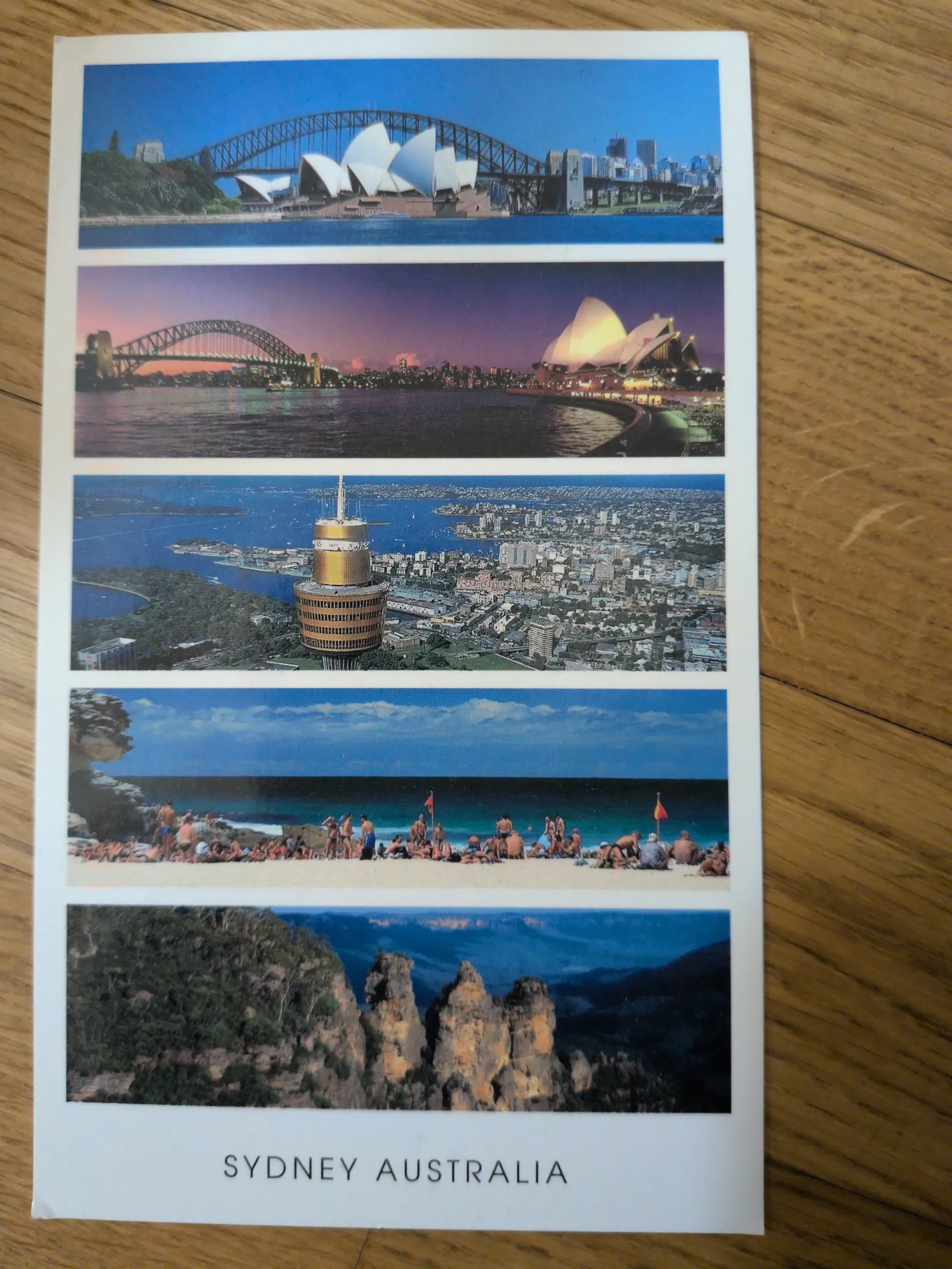 Dieses Bild ist eine Postkarte mit fünf horizontalen Aufnahmen von Sydney, Australien, die berühmte Wahrzeichen wie die Oper und Hafenbrücke, Stadtansichten, einen Strand und Naturlandschaften zeigen.