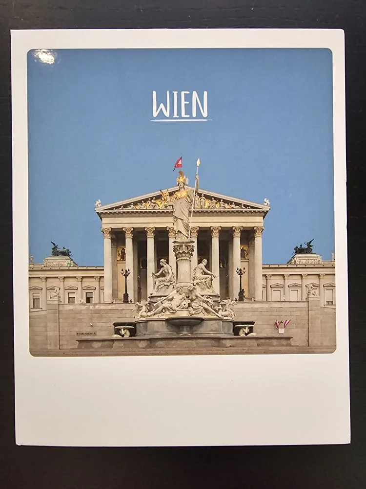 Das Bild zeigt das Wiener Parlament mit dem Pallas-Athene-Brunnen davor, unter blauem Himmel und dem Schriftzug „WIEN“.