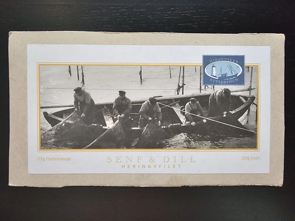 Eine beige Lebensmittelverpackung zeigt ein Schwarz-Weiß-Foto von Fischern in Booten beim Netzeinholen und trägt die Aufschrift "Senf & Dill Heringsfilet".