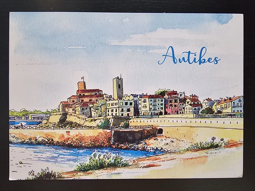Ein farbenfrohes Aquarell zeigt die Küstenstadt Antibes mit ihren historischen Gebäuden, einer Festung und einem Kirchturm am blauen Meer, darüber steht in blauer Schrift "Antibes".