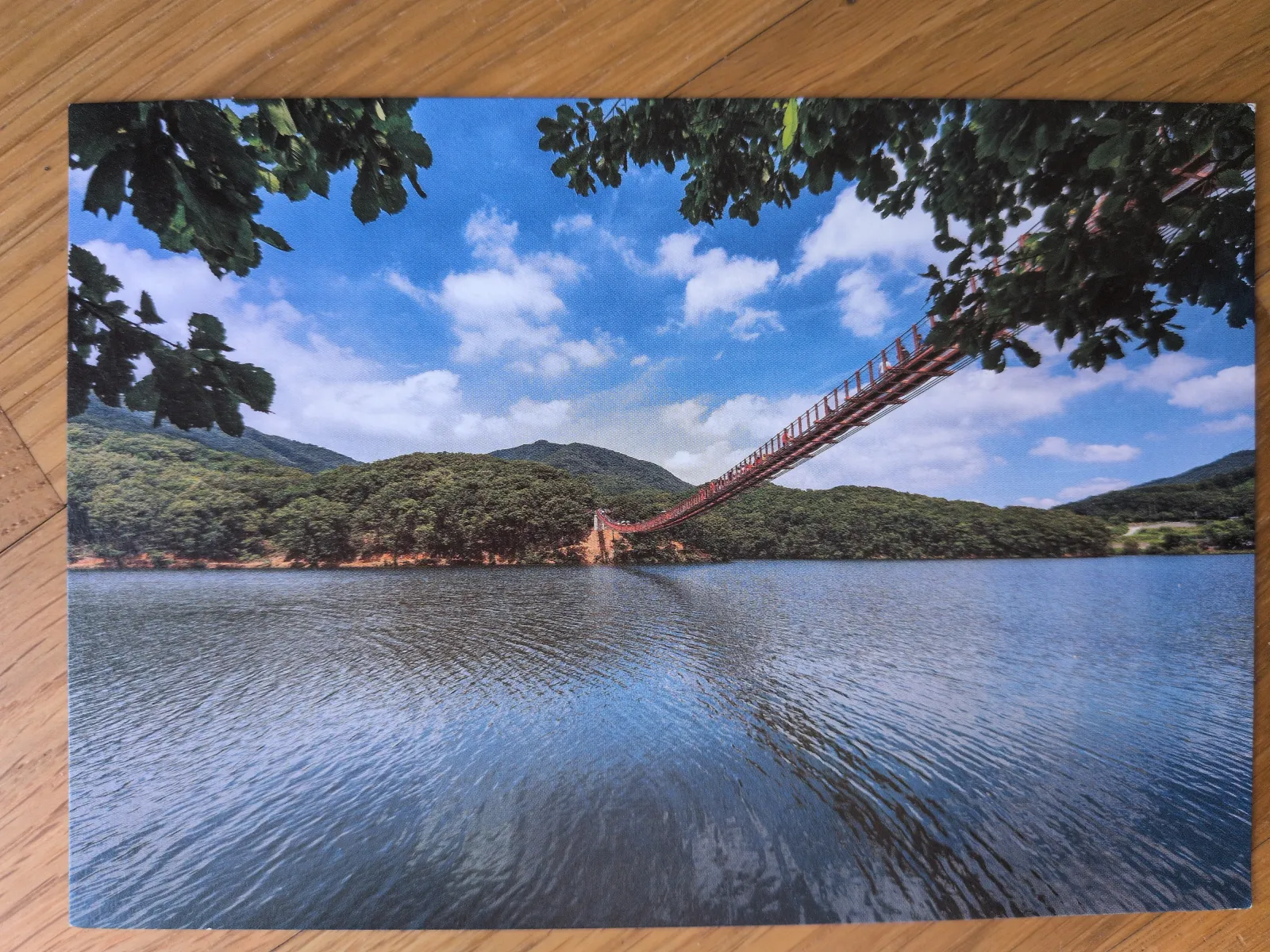 Eine rote Hängebrücke überspannt einen See, der von grünen bewaldeten Bergen unter blauem Himmel umgeben ist.