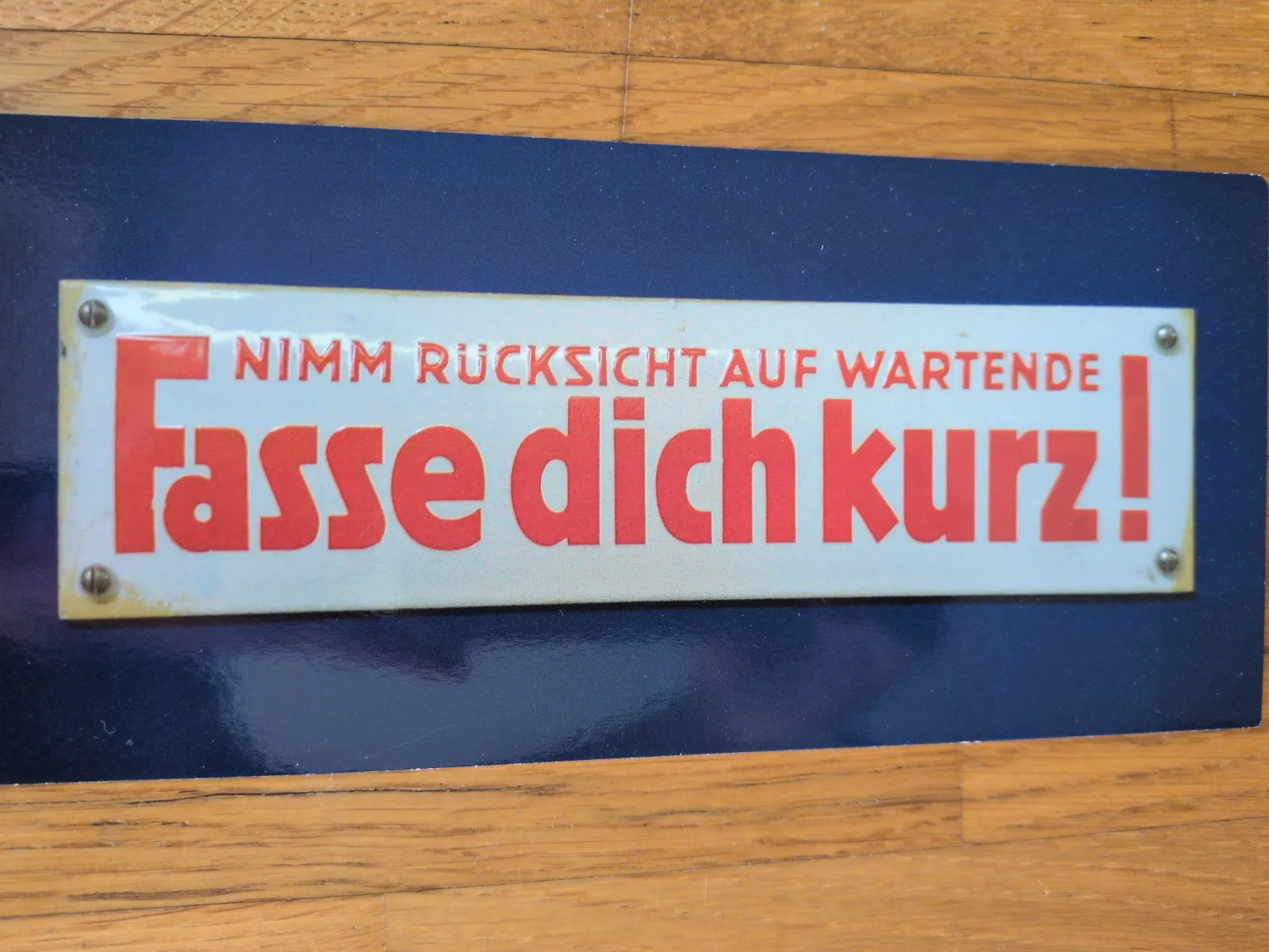 Ein weißes Schild mit roter Schrift auf blauem Untergrund zeigt die Aufforderung: "Nimm Rücksicht auf Wartende, Fasse dich kurz!".