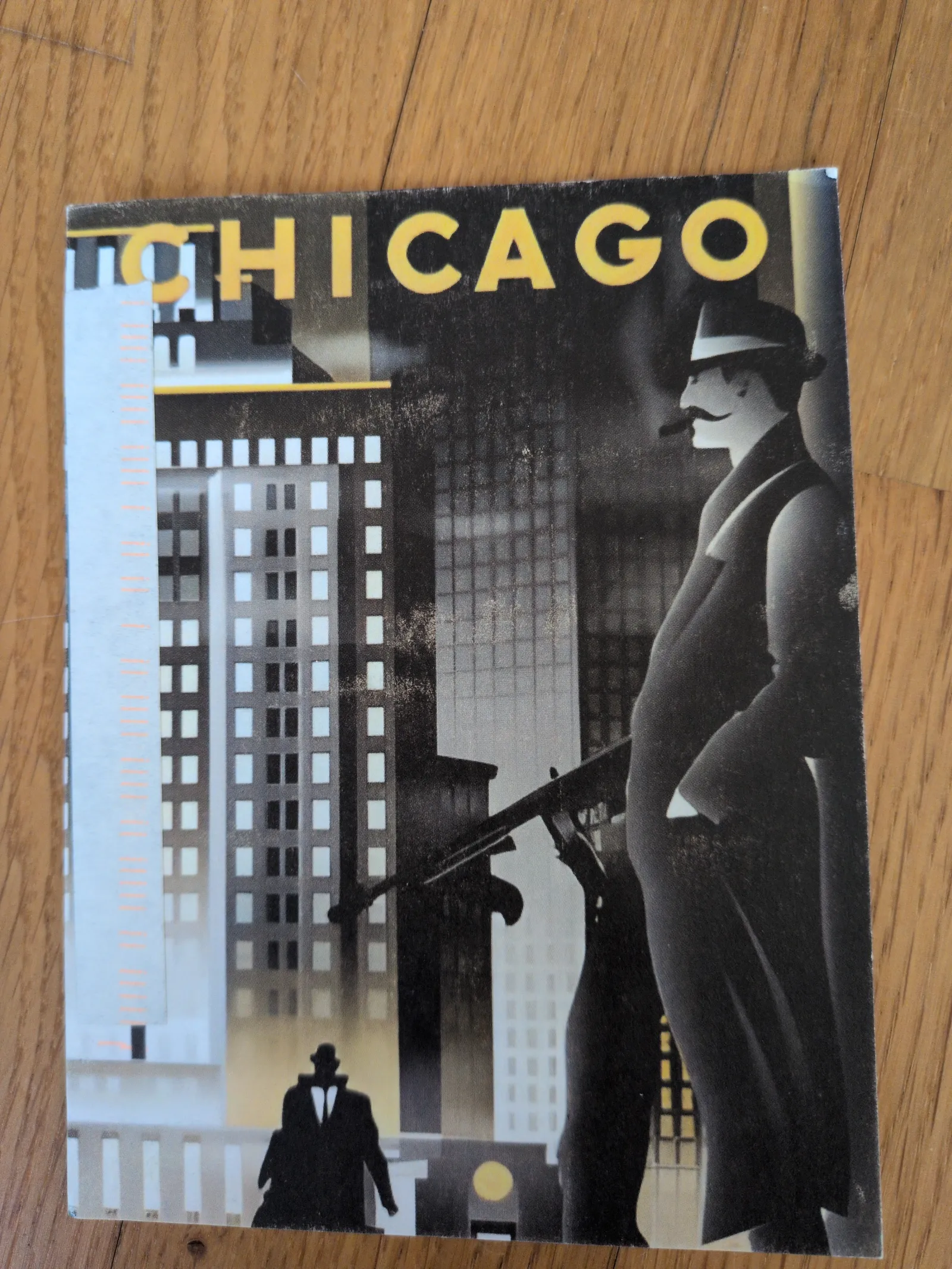Ein Art-déco-Plakat mit dem Schriftzug „CHICAGO“ zeigt einen Mann in Hut und Mantel mit einer Maschinenpistole vor einer dunklen Stadtkulisse.